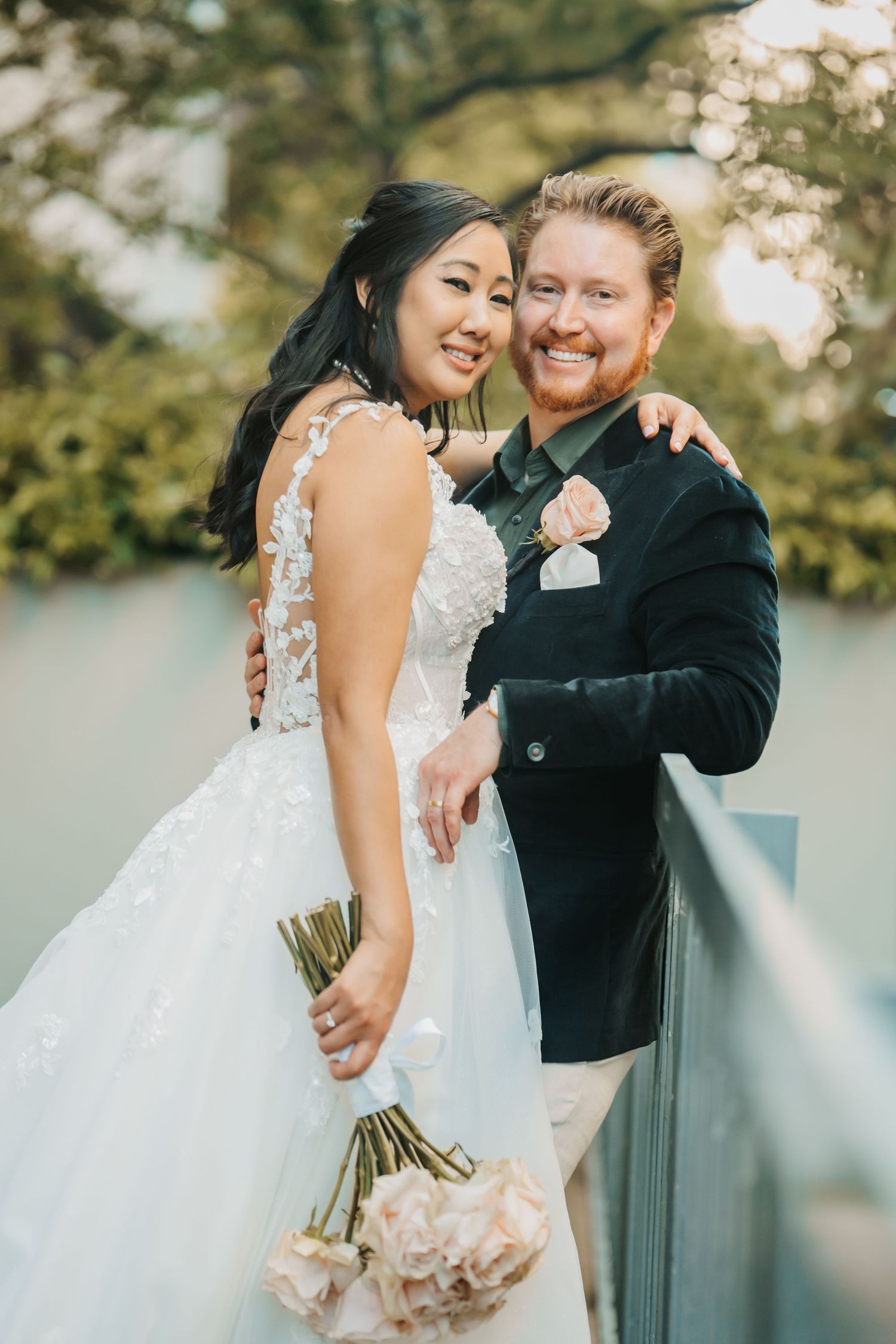Wedding couple shares tender moment on bridge holding pink bouquet in scenic outdoor setting during sunset.