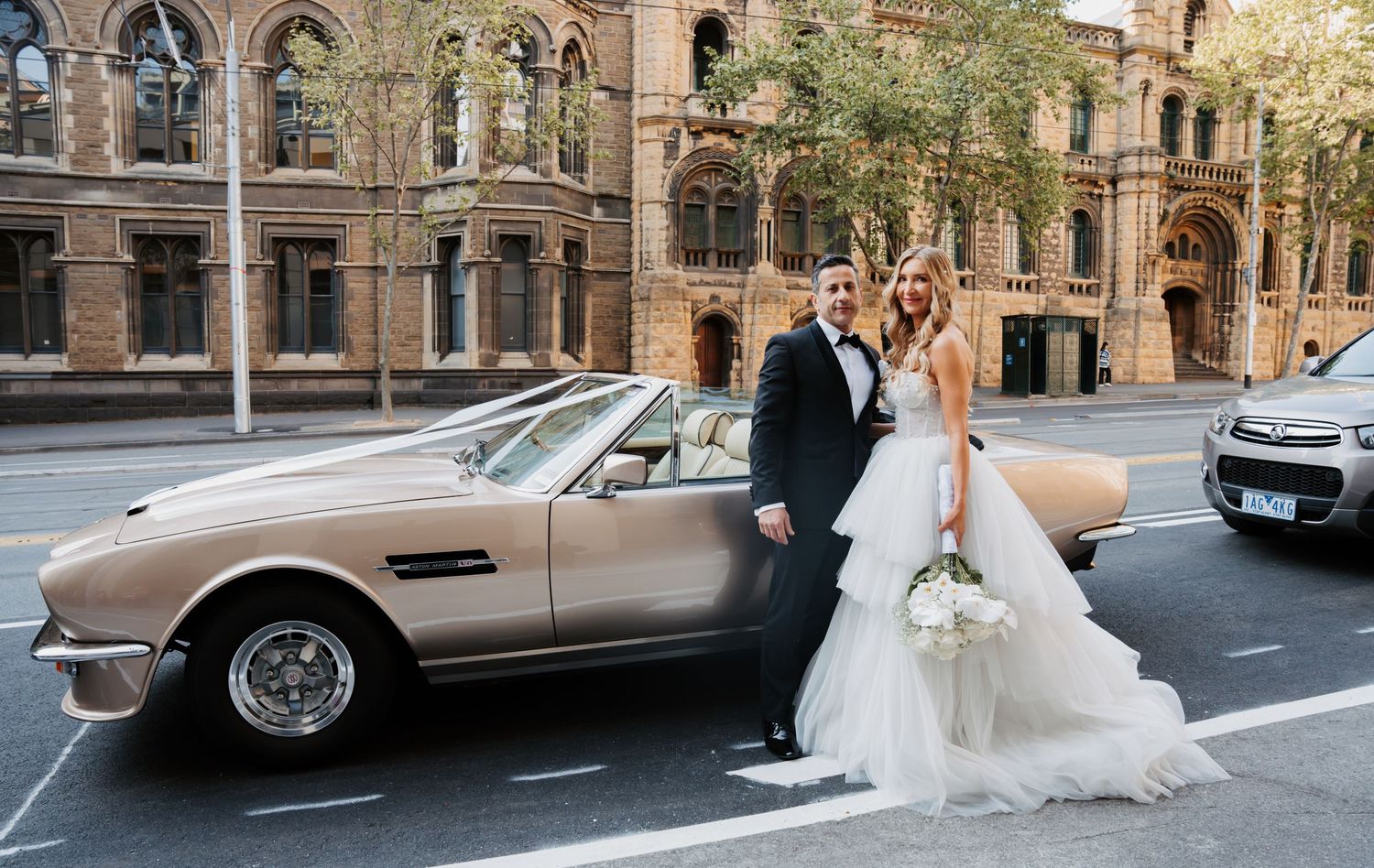 Vintage Aston Martin parked on city street outside historic Gothic architecture building with wedding couple nearby.