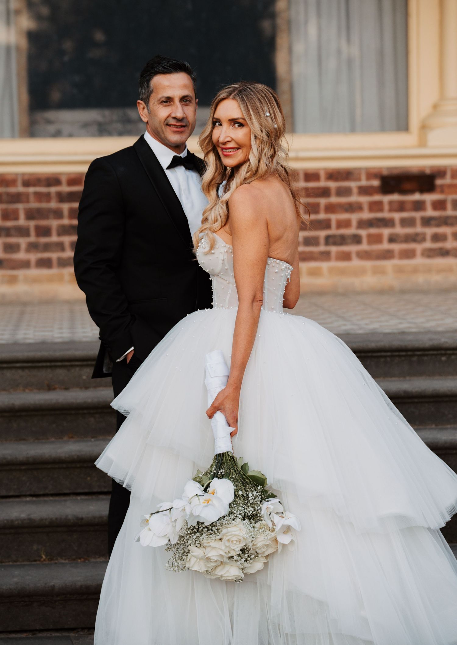 A romantic wedding couple embraces on stone steps, with the bride wearing a layered tulle ballgown and holding white flowers.