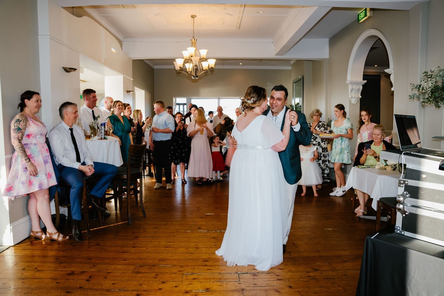 A couple shares their first dance at a wedding reception while guests watch from the sides of a hardwood-floored venue.