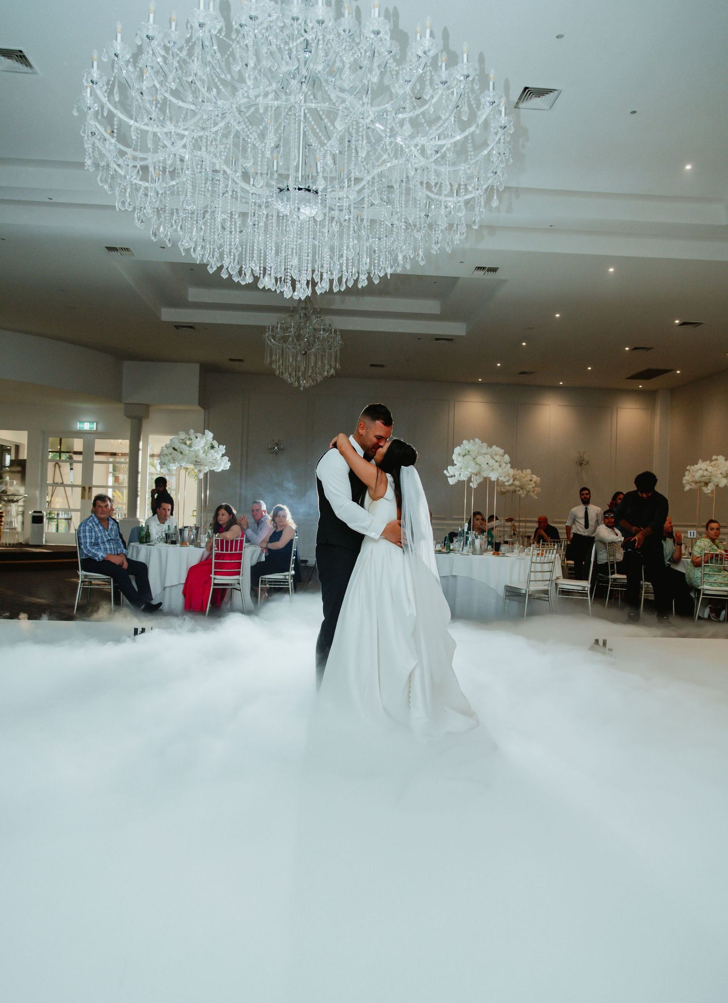 A couple shares their first dance under a crystal chandelier with fog effects on the dance floor in an elegant reception hall.