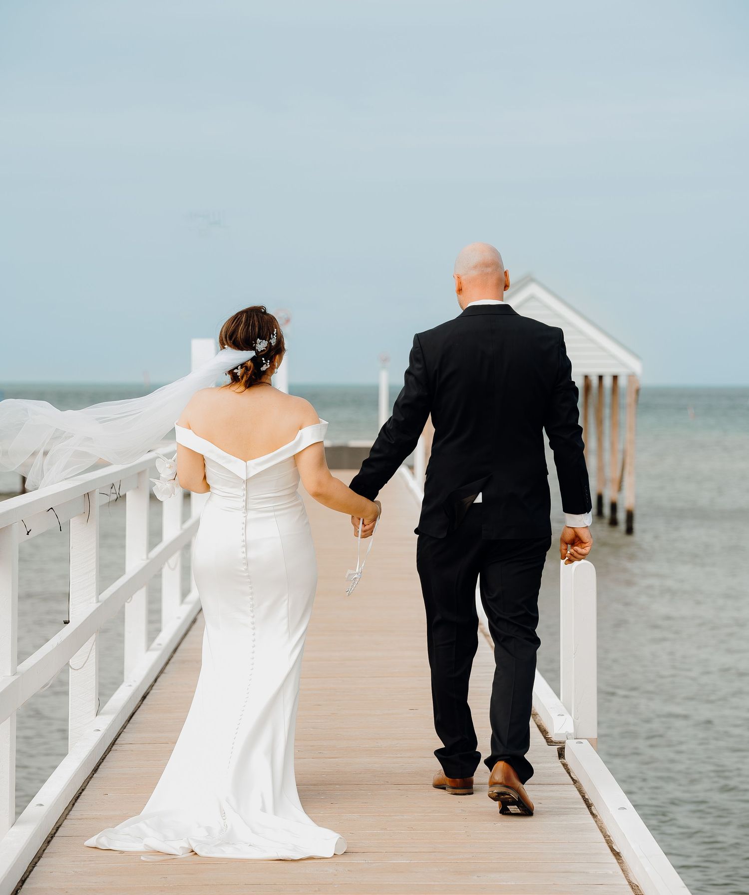 A couple in wedding attire walks along a white wooden pier overlooking the ocean.