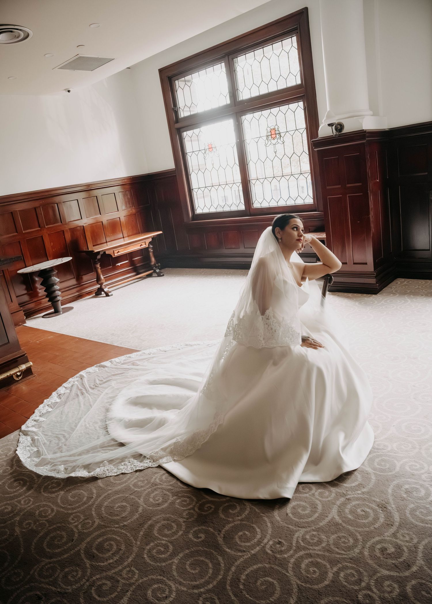 Bride in a white ball gown with long veil sits gracefully in an elegant wood-paneled room with stained glass windows.