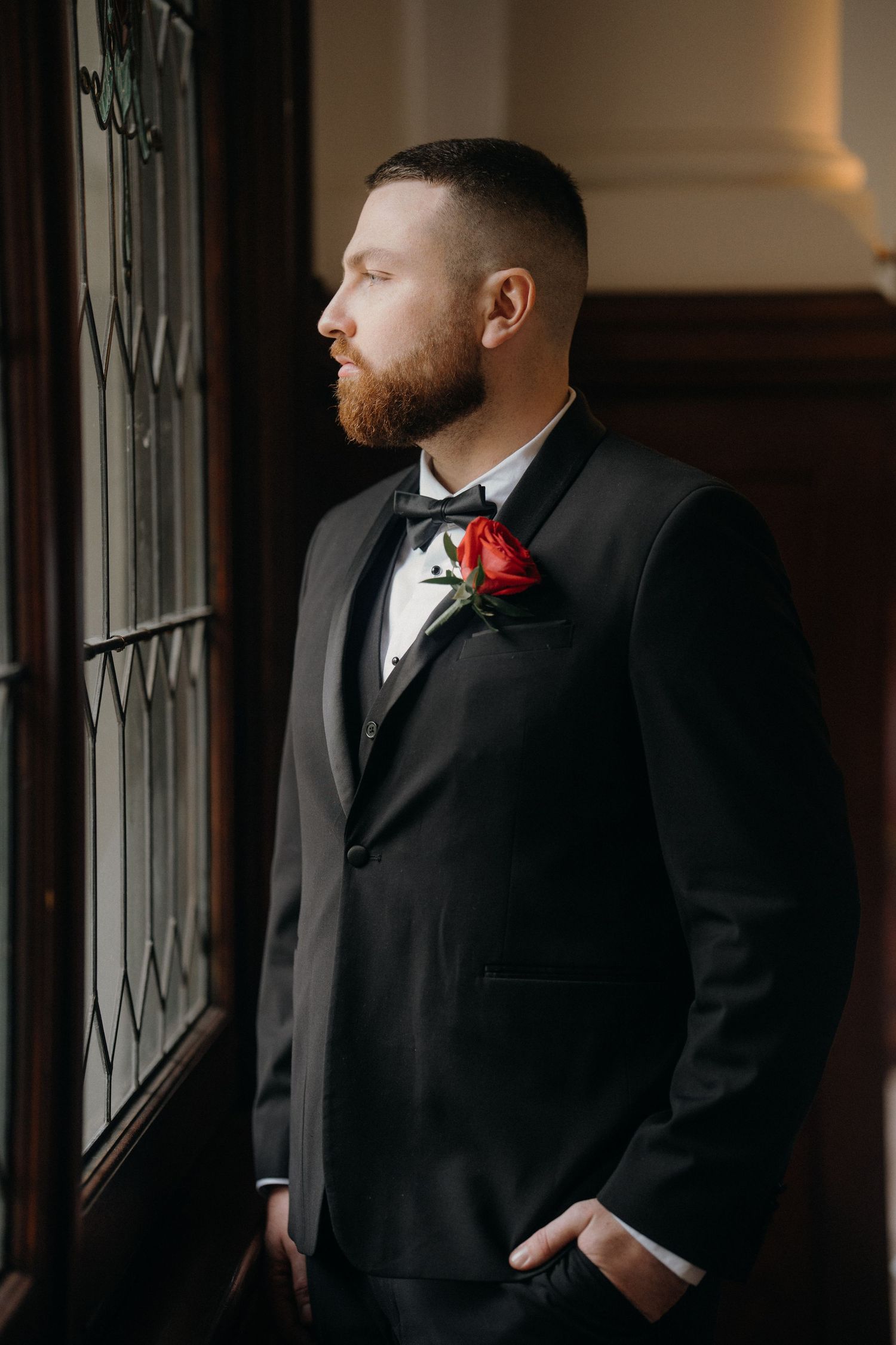 Groom in black tuxedo with red rose boutonniere and bow tie stands by window in elegant venue.