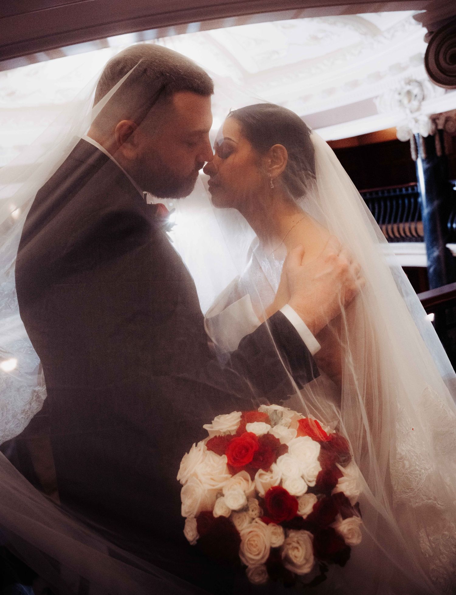 Bride and groom sharing a romantic kiss under a veil, bride holding a red and white rose bouquet on their wedding day.