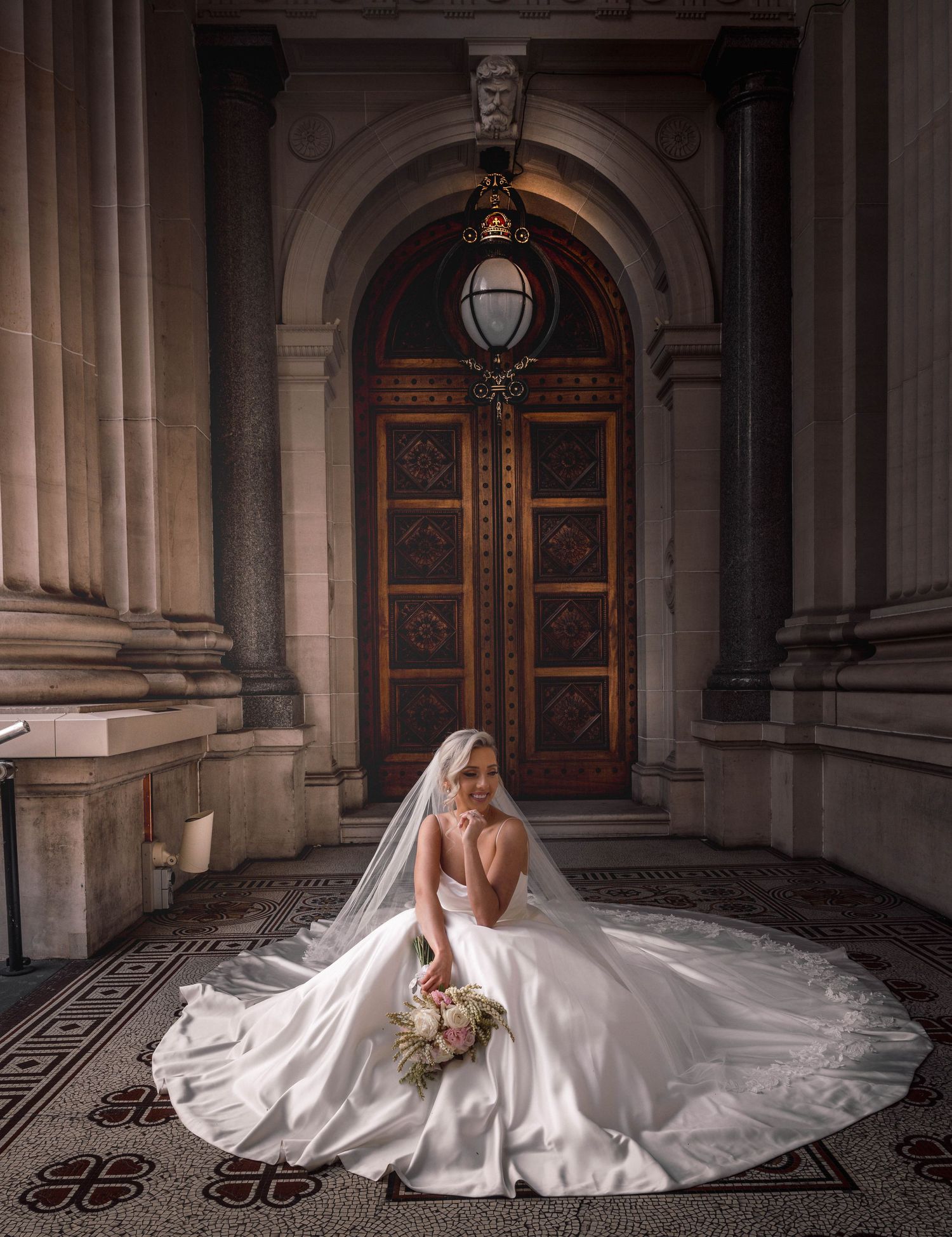 Bride in flowing white gown poses elegantly on ornate marble floor against backdrop of grand arched doorways.