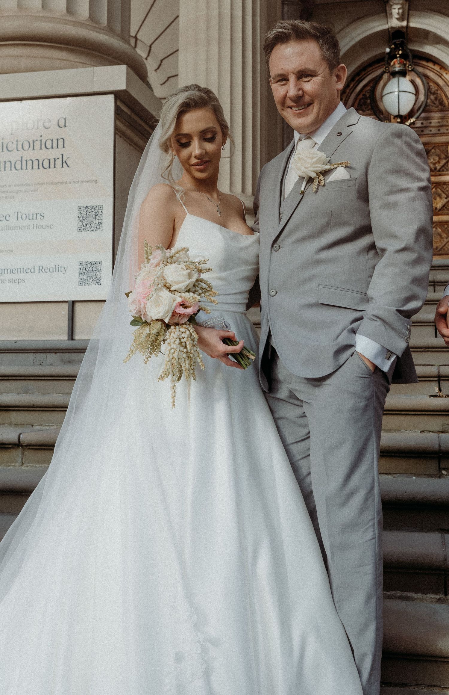 Newlywed couple in grey suit and white wedding dress pose together in front of historic building entrance.