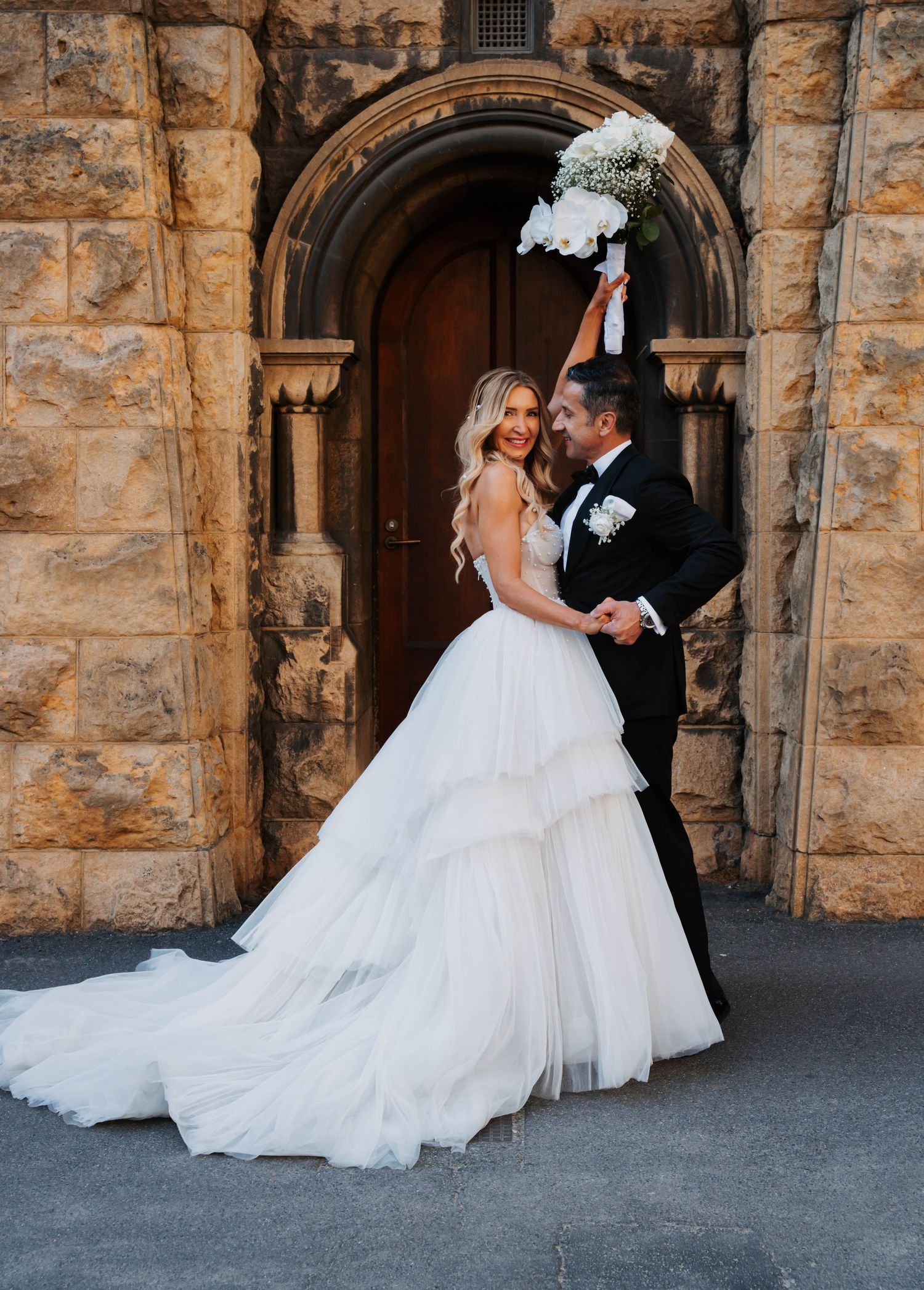 A romantic wedding photo of a couple in front of a stone archway, with the bride in a flowing tulle gown holding a bouquet.