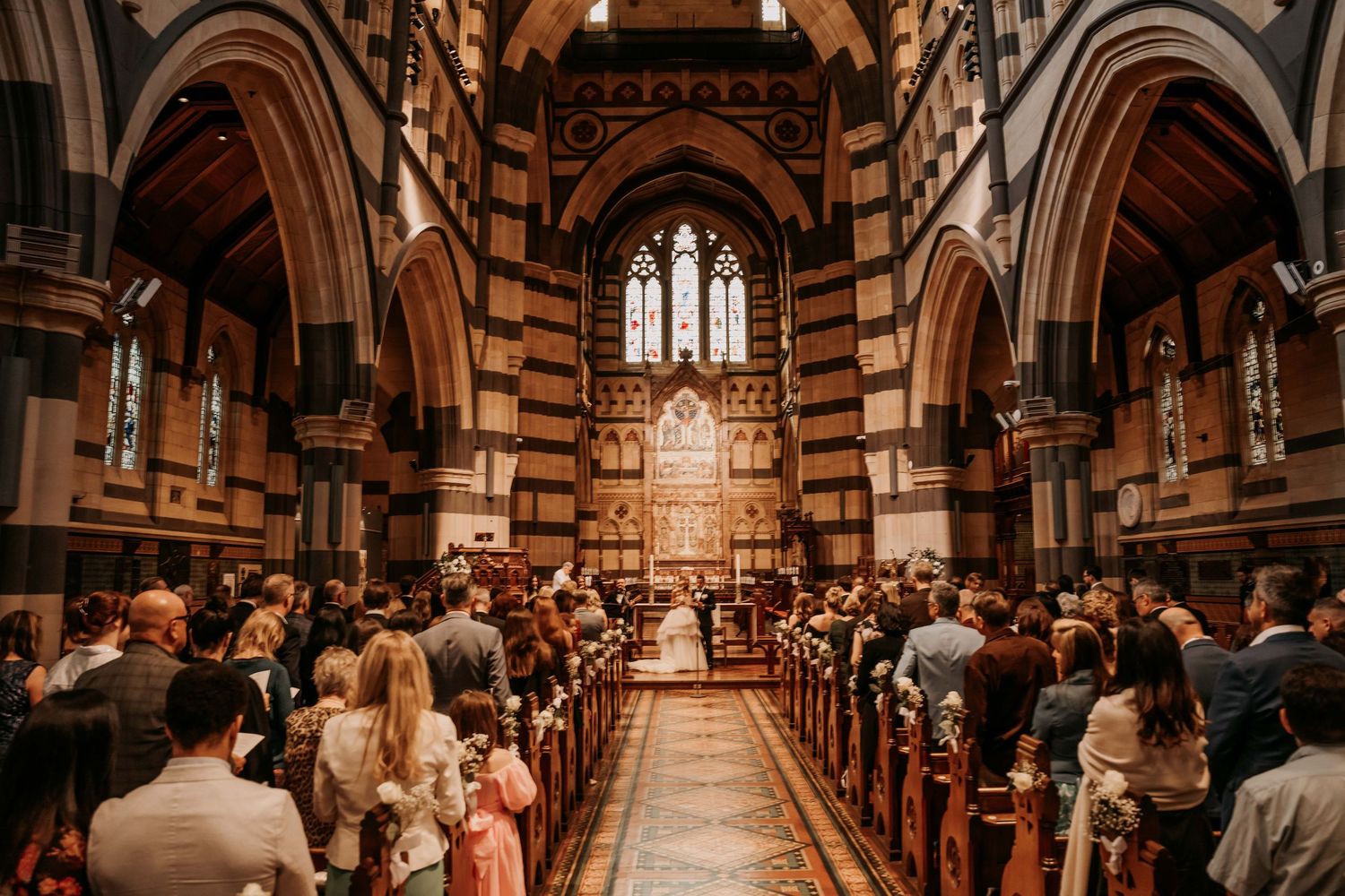 Guests gather inside a grand Gothic church with vaulted ceilings and wooden pews during a wedding ceremony.