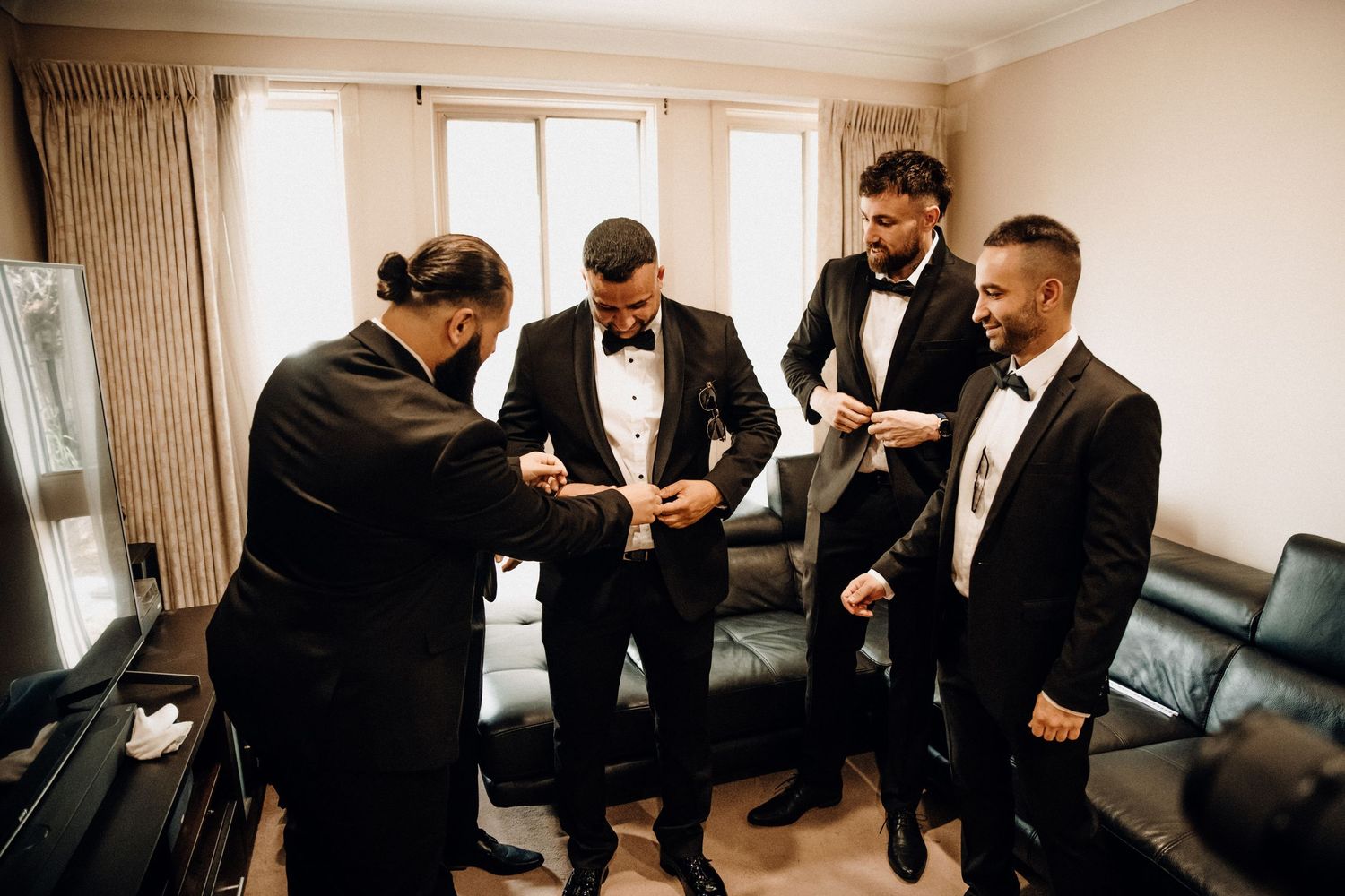 A group of people in formal black tuxedos gather together in a hotel room before an event.