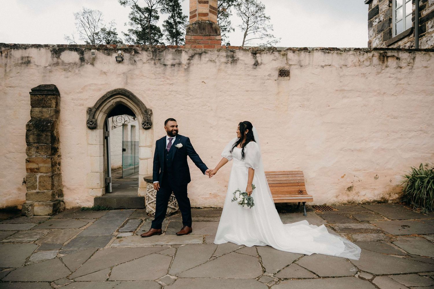 A couple holds hands against a rustic stone wall during their wedding ceremony, with the bride wearing a flowing white gown.