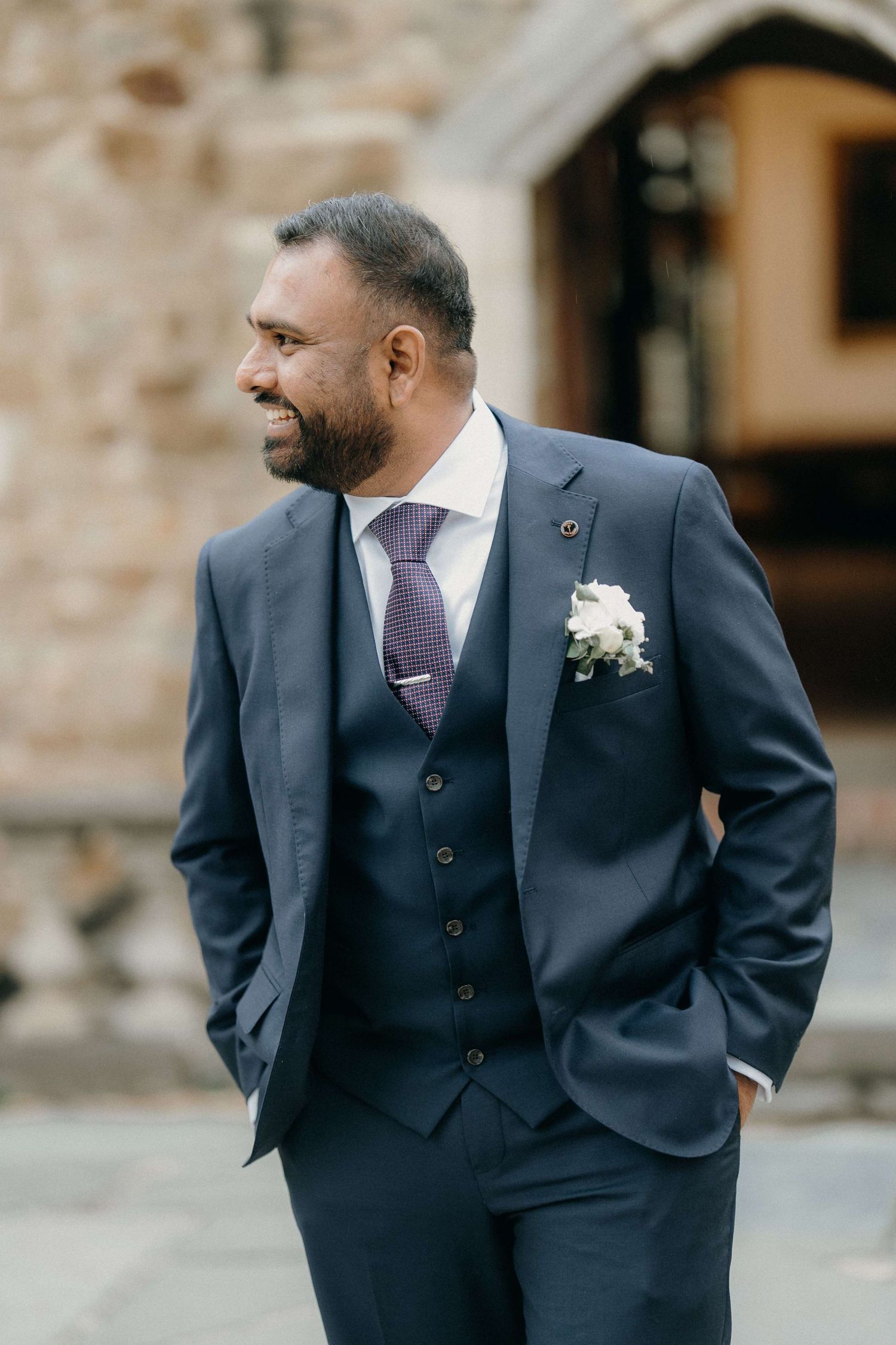 A groom in a stylish navy three-piece suit with purple tie and white boutonniere smiles while standing outdoors.