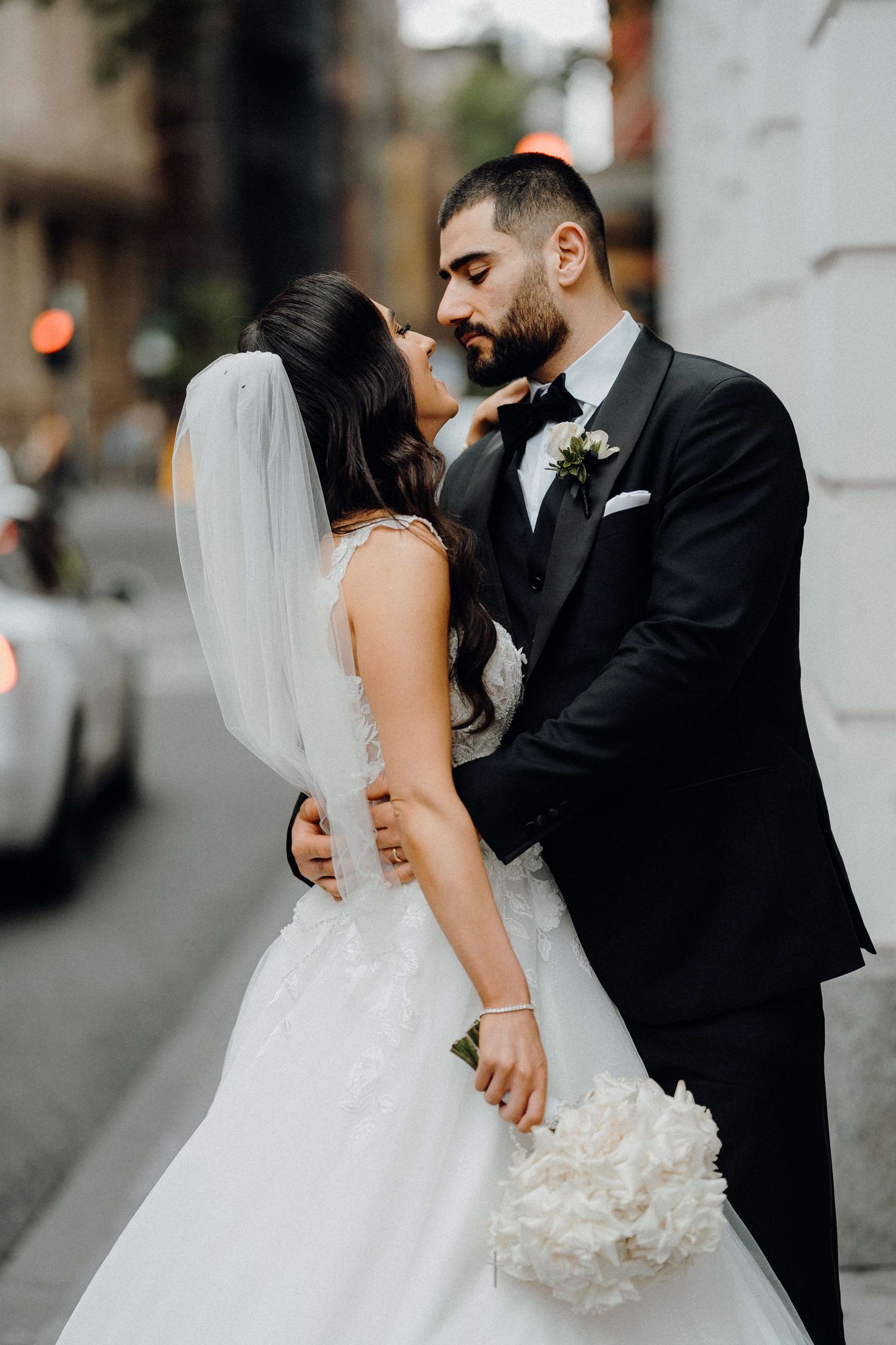 A romantic newlywed couple embraces on their wedding day, with the bride in white holding a cream bouquet.