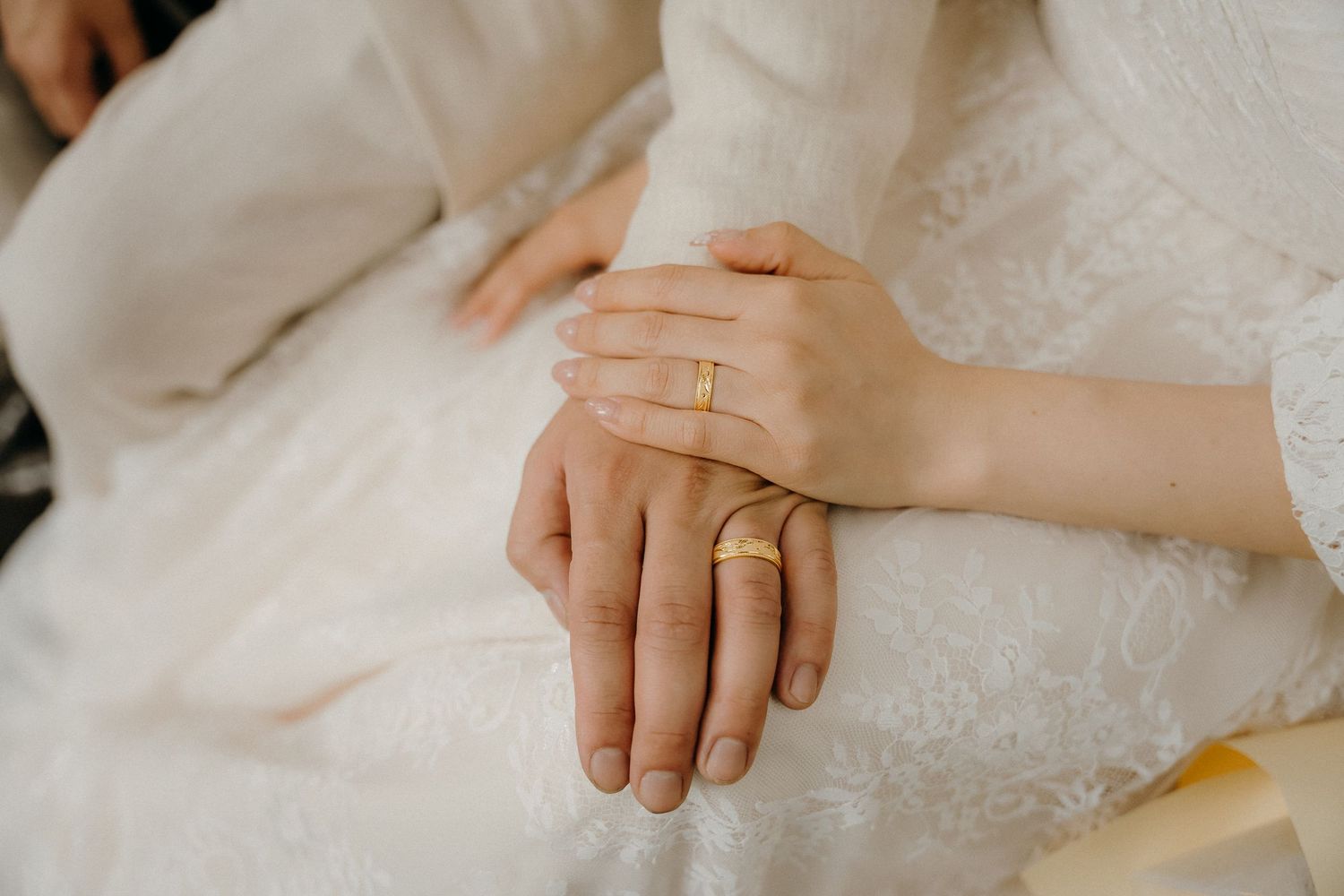 Close-up view of hands wearing wedding rings against white lace fabric background.