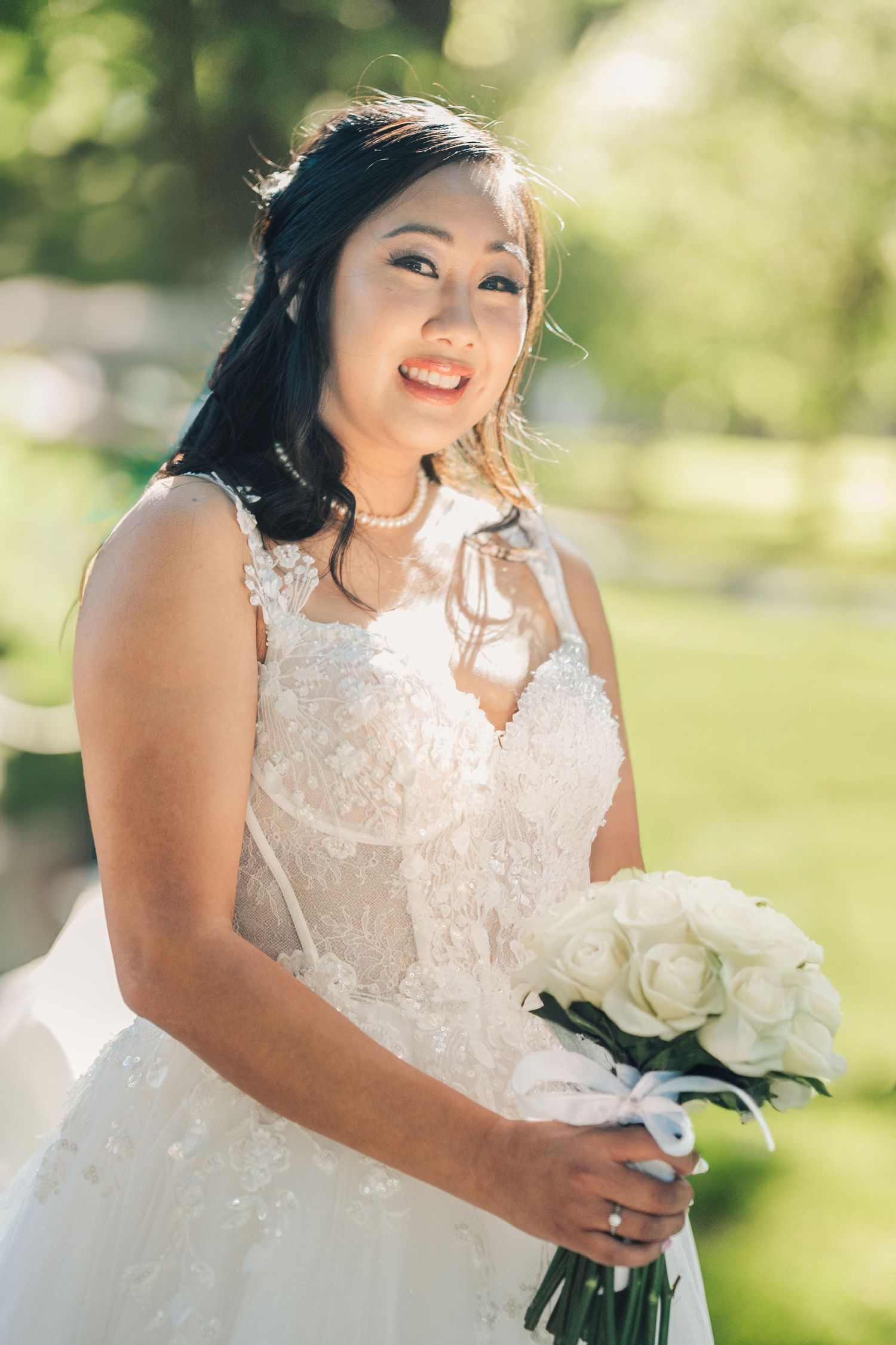 A joyful bride in a lace white dress holds a bouquet of white roses while posing for outdoor wedding photos.