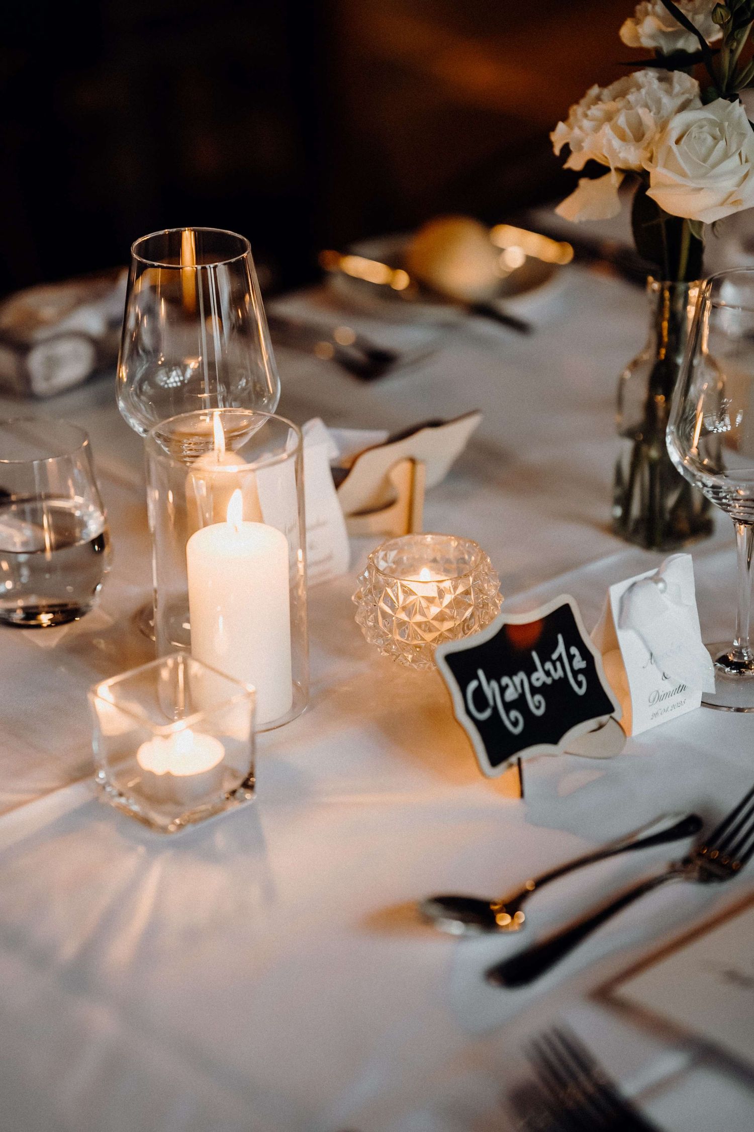 Elegant wedding table setting with glowing candles, crystal glassware, white linens and a small chalkboard name card.