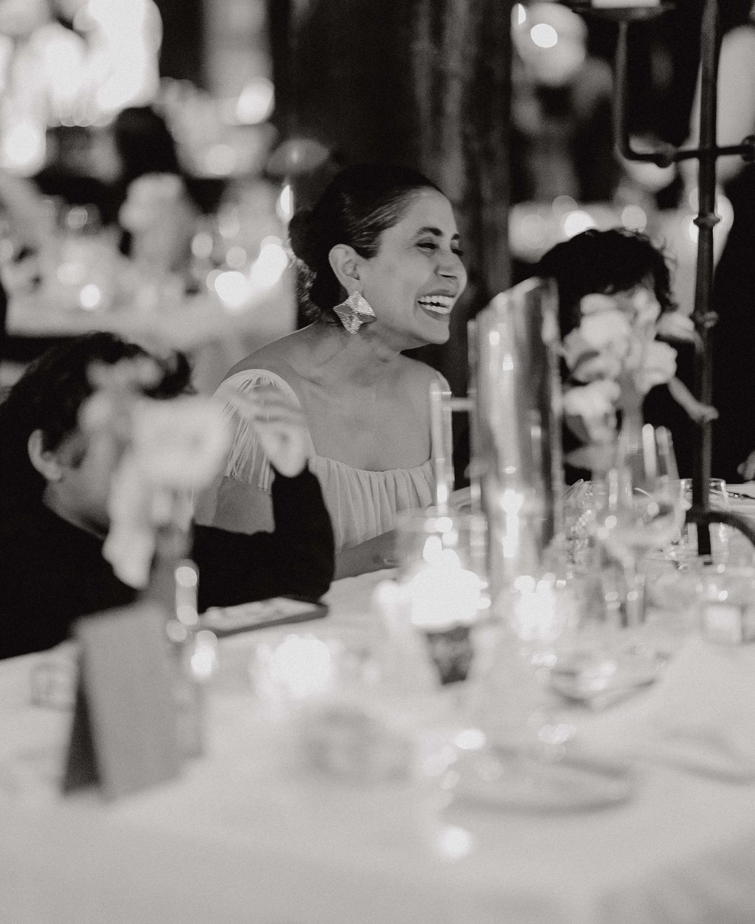 Wedding guests laugh and celebrate at elegant candlelit reception dinner with white floral centerpieces.