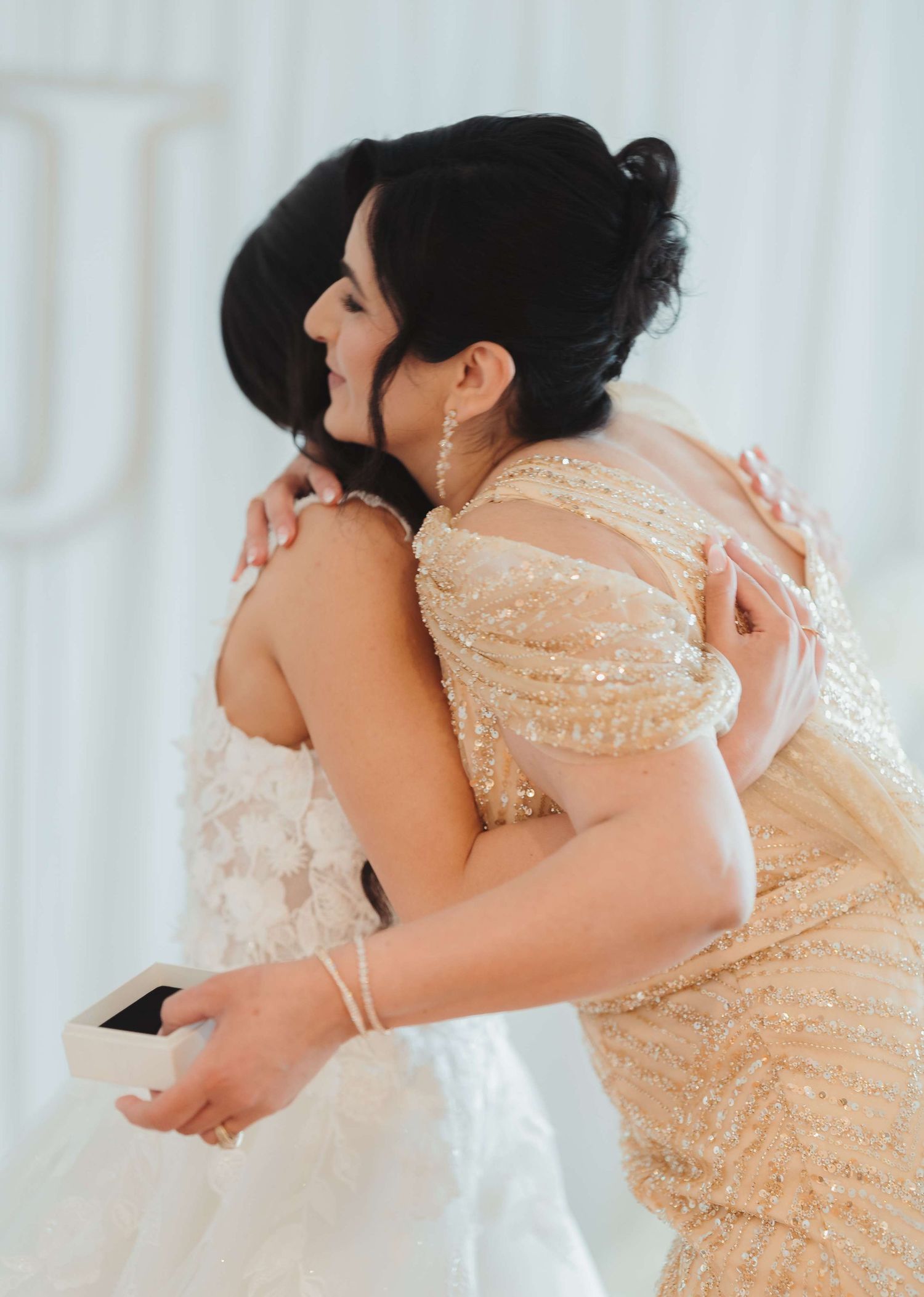 Two people share an emotional embrace at a wedding, one wearing a sparkly champagne-colored dress and the other in white.
