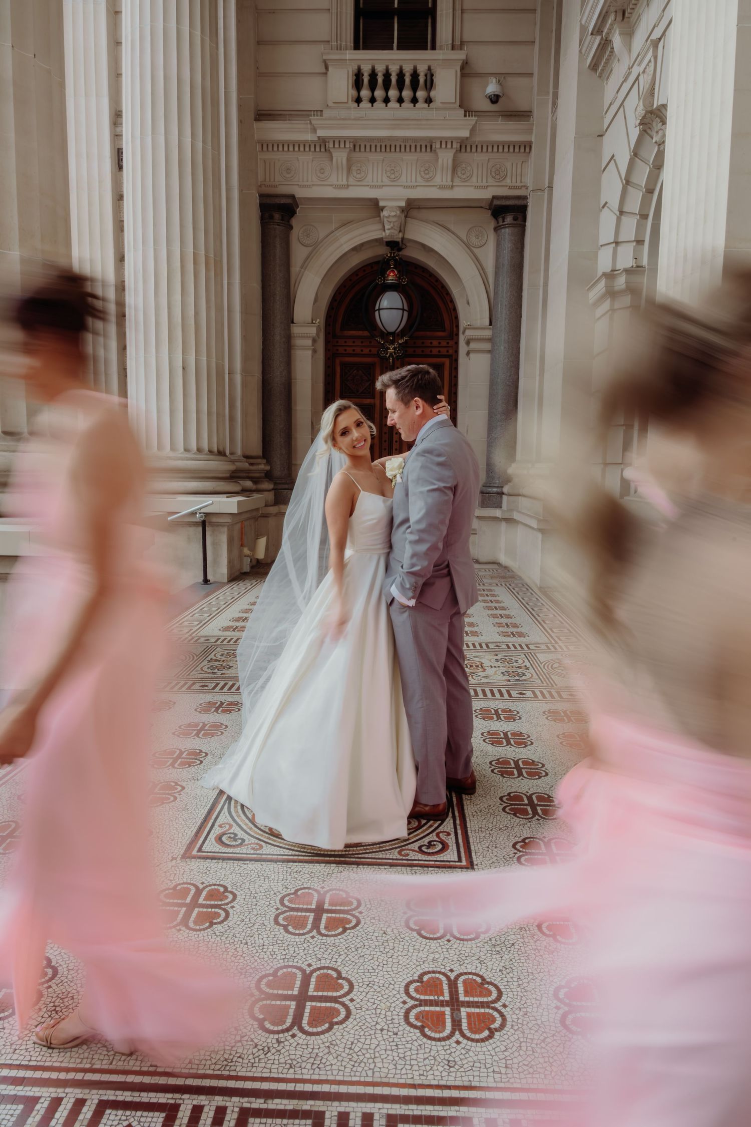 A couple embraces in an elegant marble hallway with ornate architectural details as pink-dressed figures blur past.