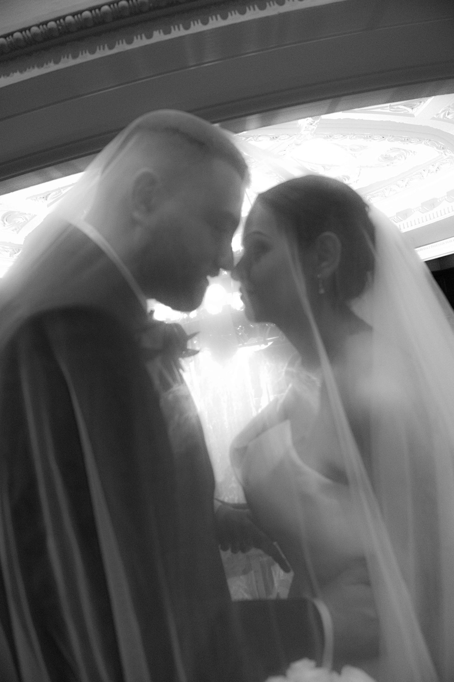 Black and white photo of bride and groom touching foreheads under wedding veil.