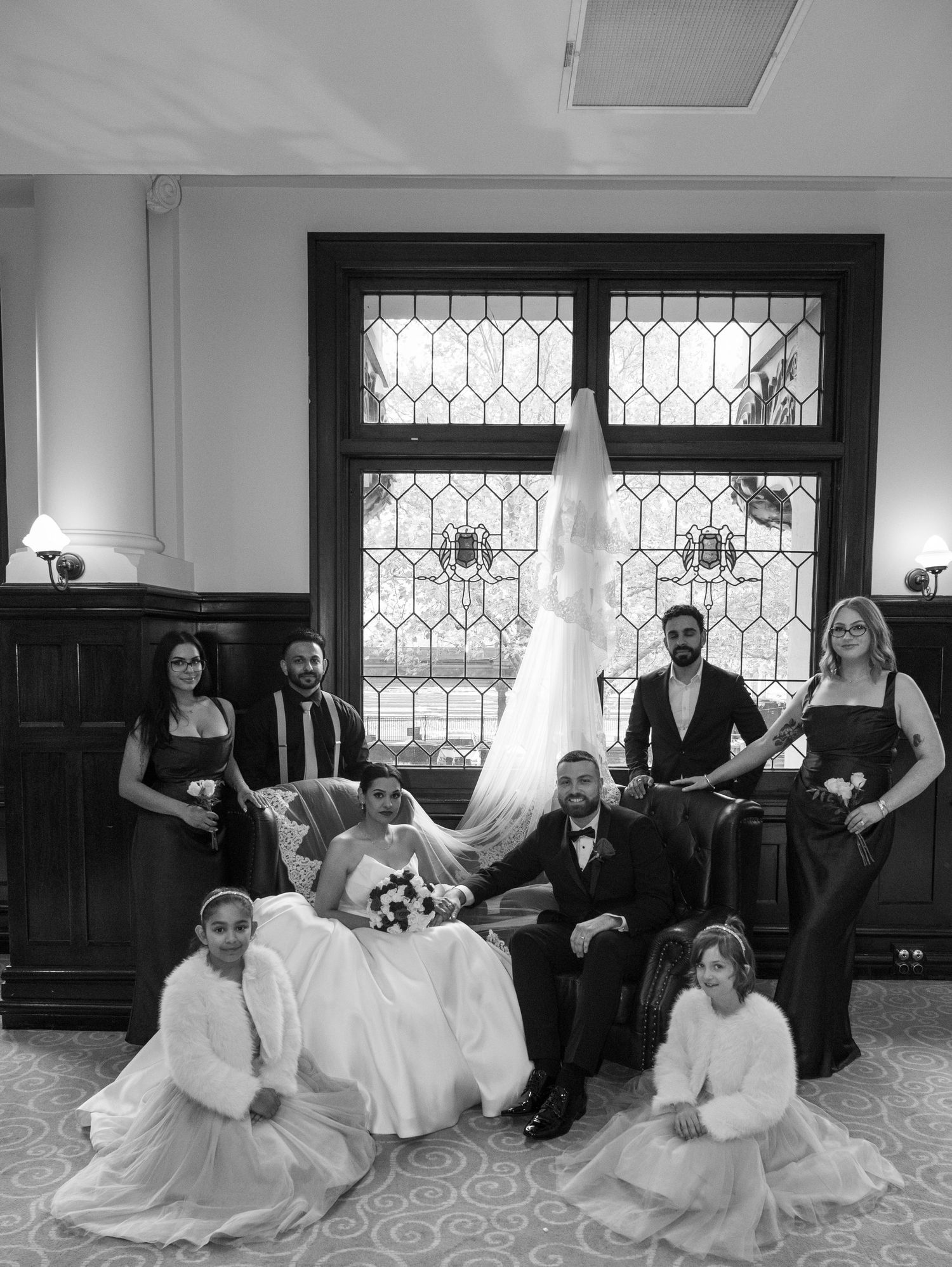 Black and white wedding party photo with bride, groom, bridesmaids, groomsmen, and flower girls posed near a stained glass window.