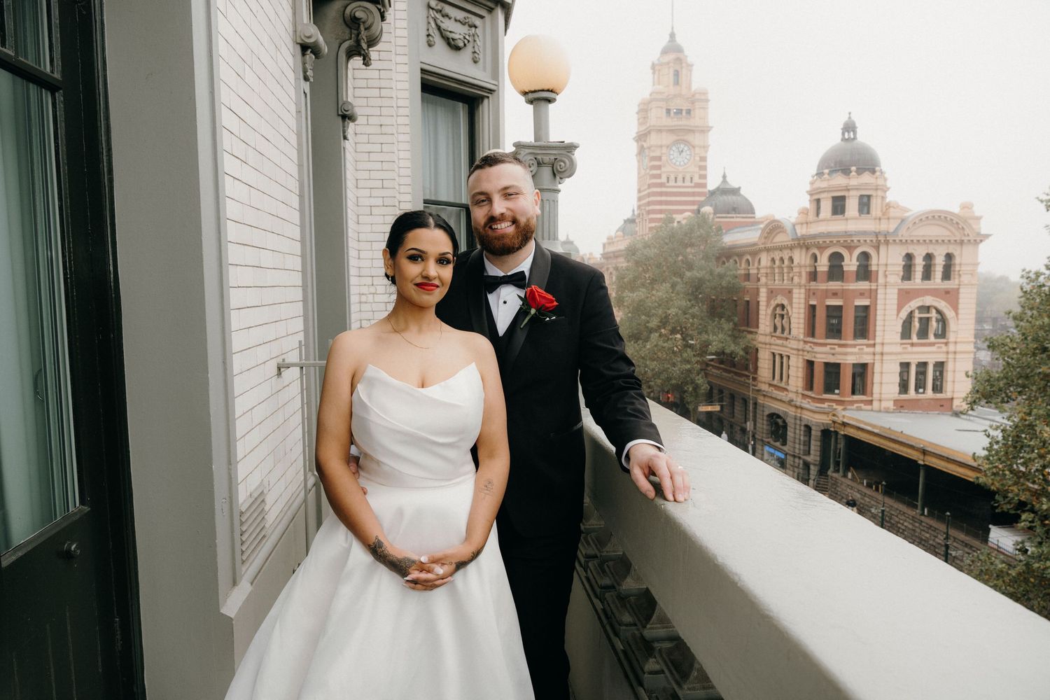 Bride and groom posing on a balcony in formal wedding attire with historic city buildings in the background.