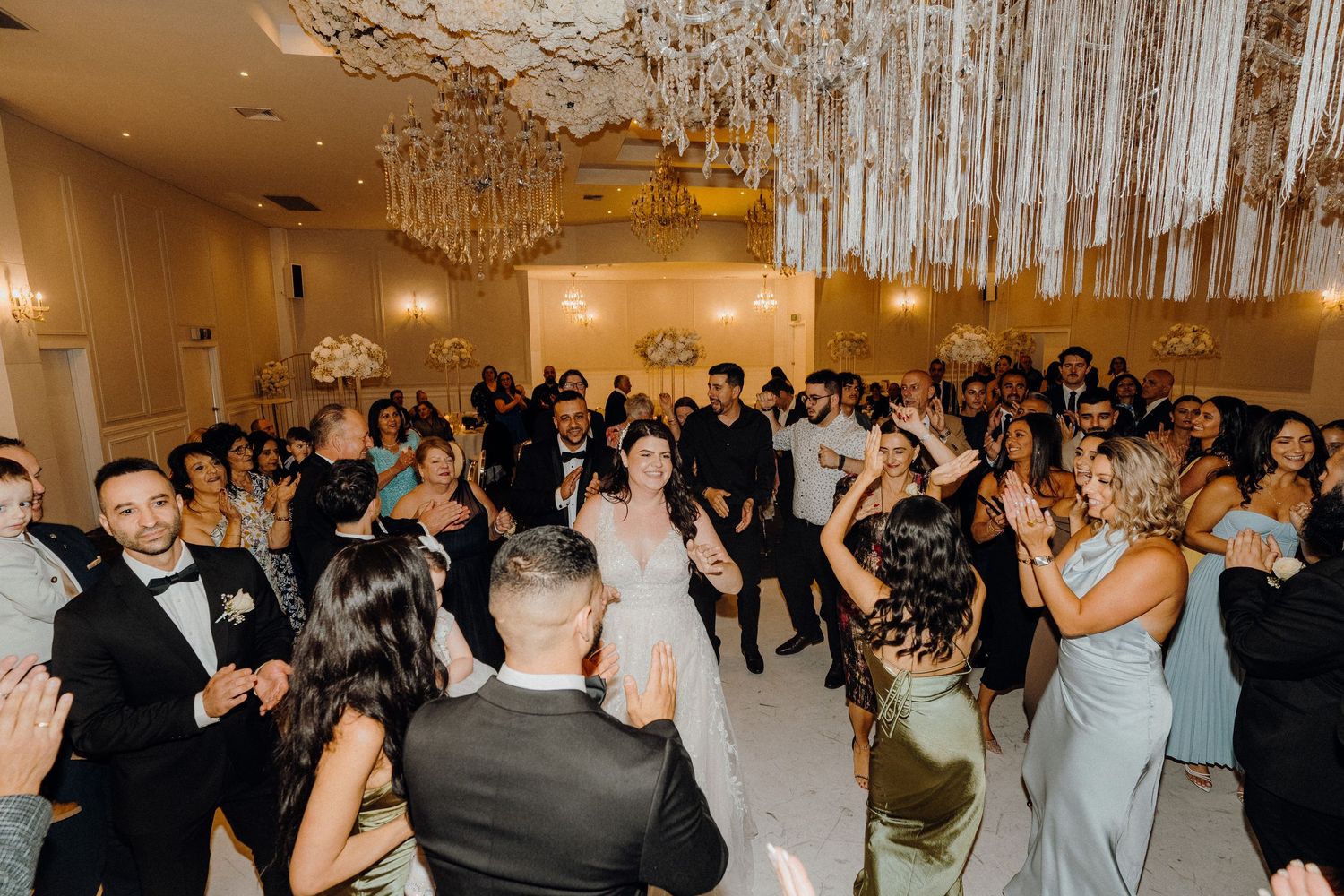 People dance in an elegant ballroom with chandeliers during a formal reception event.