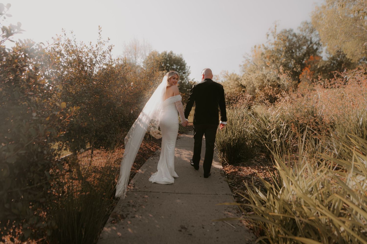 A couple in wedding attire walks down a sunlit path surrounded by autumn foliage and tall grass.