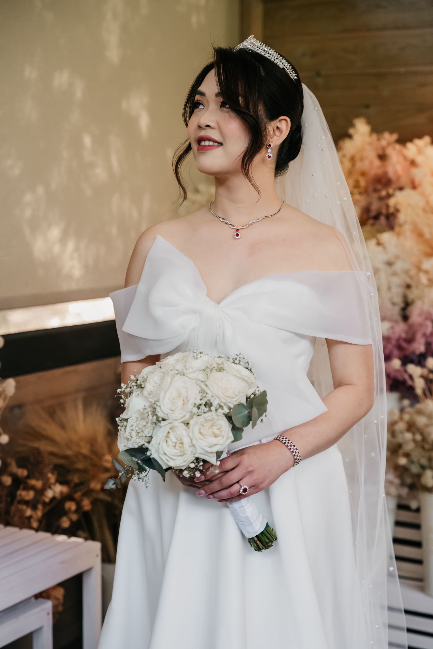 A bride in an elegant white off-shoulder wedding dress holds a beautiful white and green floral bouquet.
