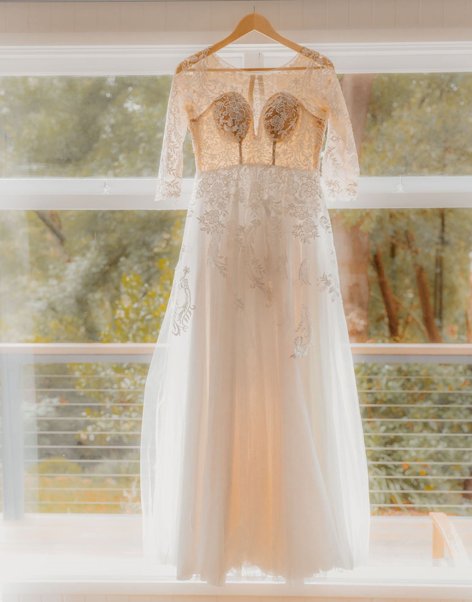 A romantic white lace wedding dress with delicate details hangs elegantly from a wooden hanger by a window.