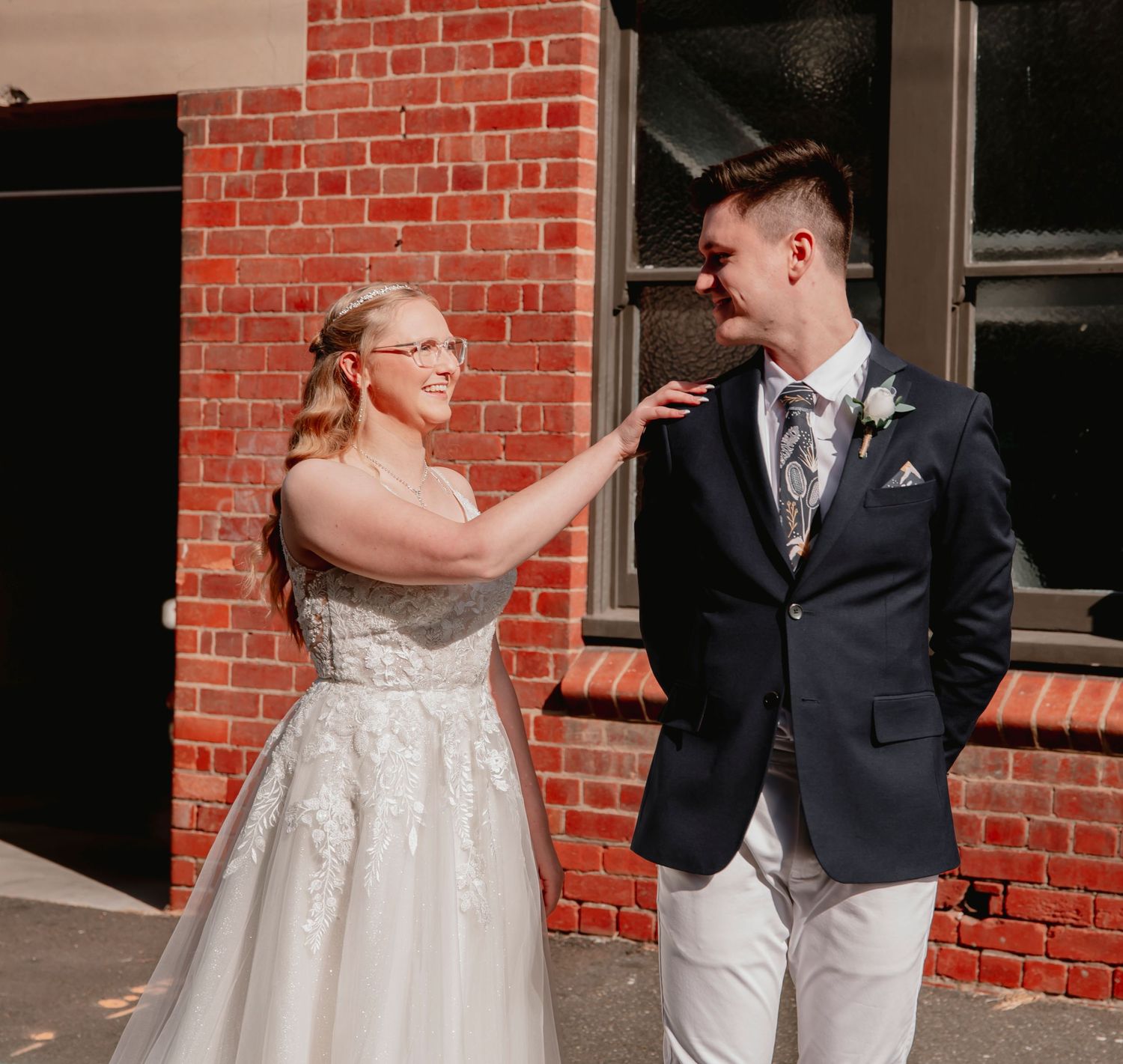 A couple shares a tender moment in front of a brick building, with the bride wearing a white gown and groom in a suit.