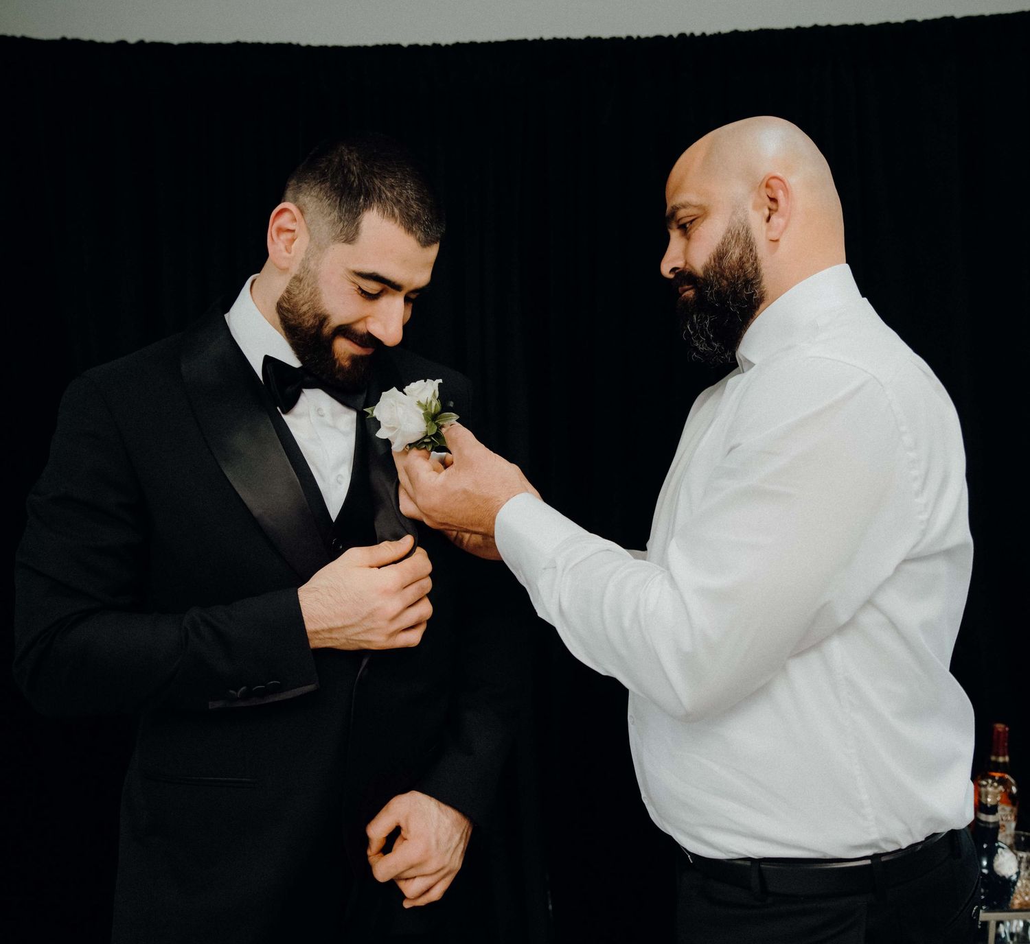 Two people sharing a happy moment during a wedding ceremony as they prepare boutonnieres on a black background.