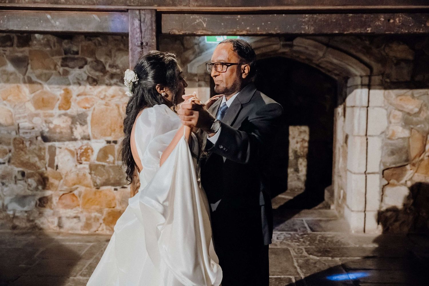 Wedding couple shares an intimate moment against a rustic stone wall backdrop in soft lighting.