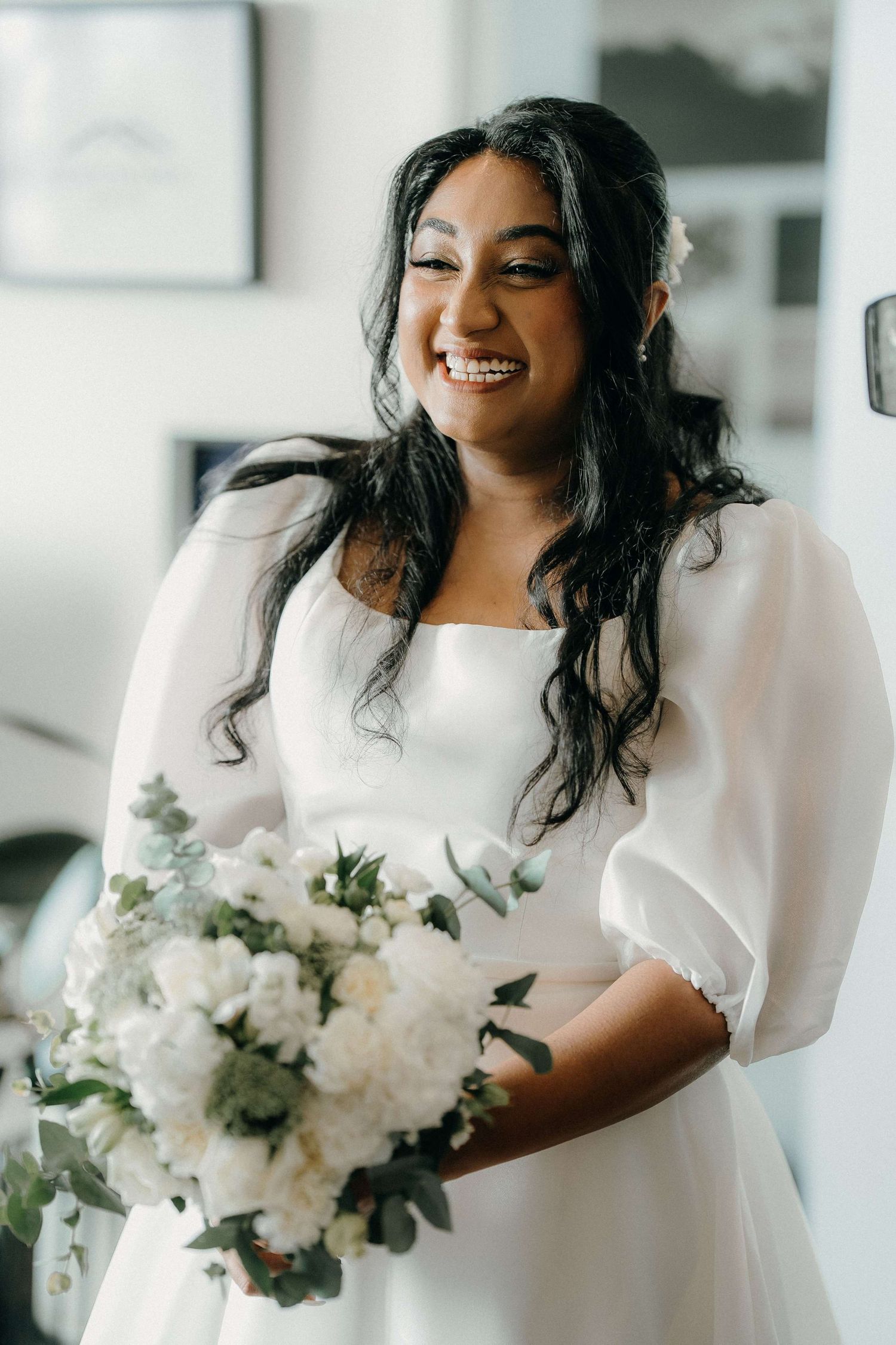 A radiant bride in a flowing white dress laughs while holding a lush bouquet of white flowers.