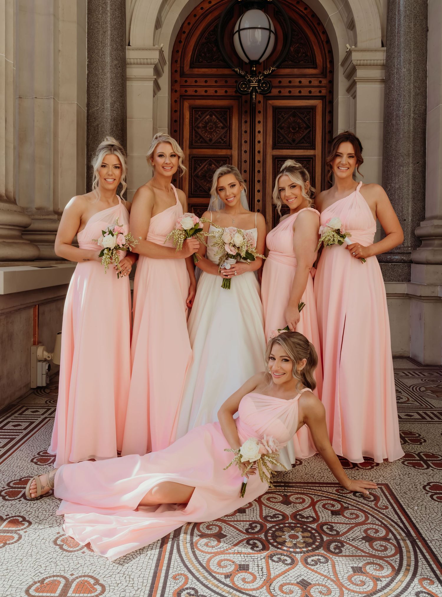 A bridal party poses in matching blush pink bridesmaid dresses and a white wedding gown on ornate tiled floor.
