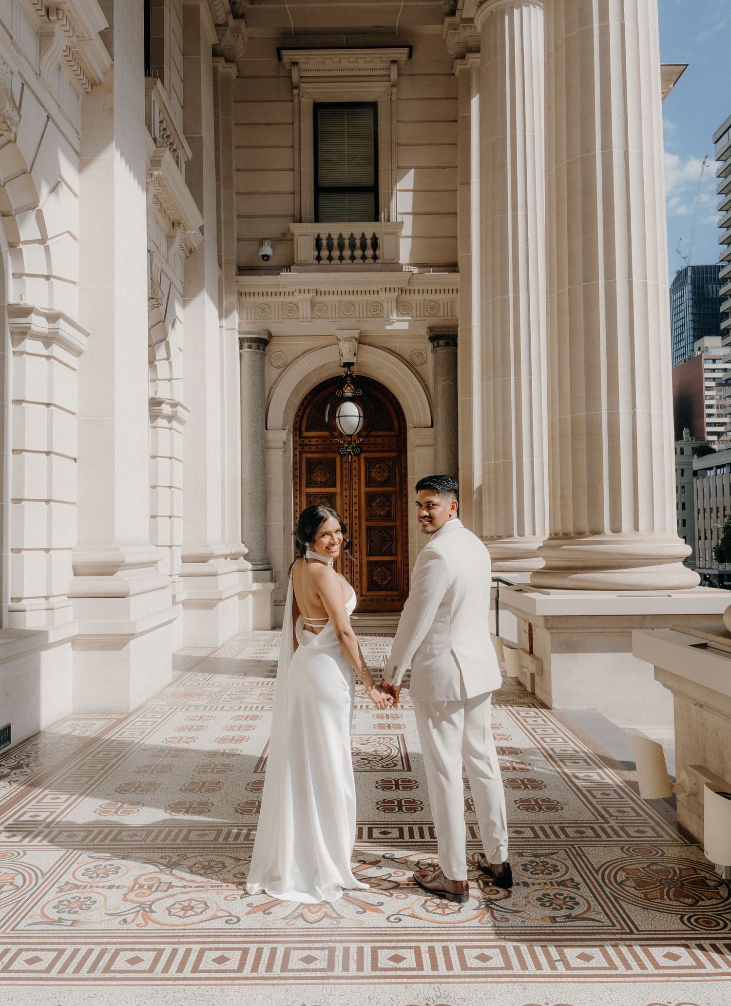Couple in elegant white outfits shares romantic moments in a grand colonnade with ornate patterned floors.