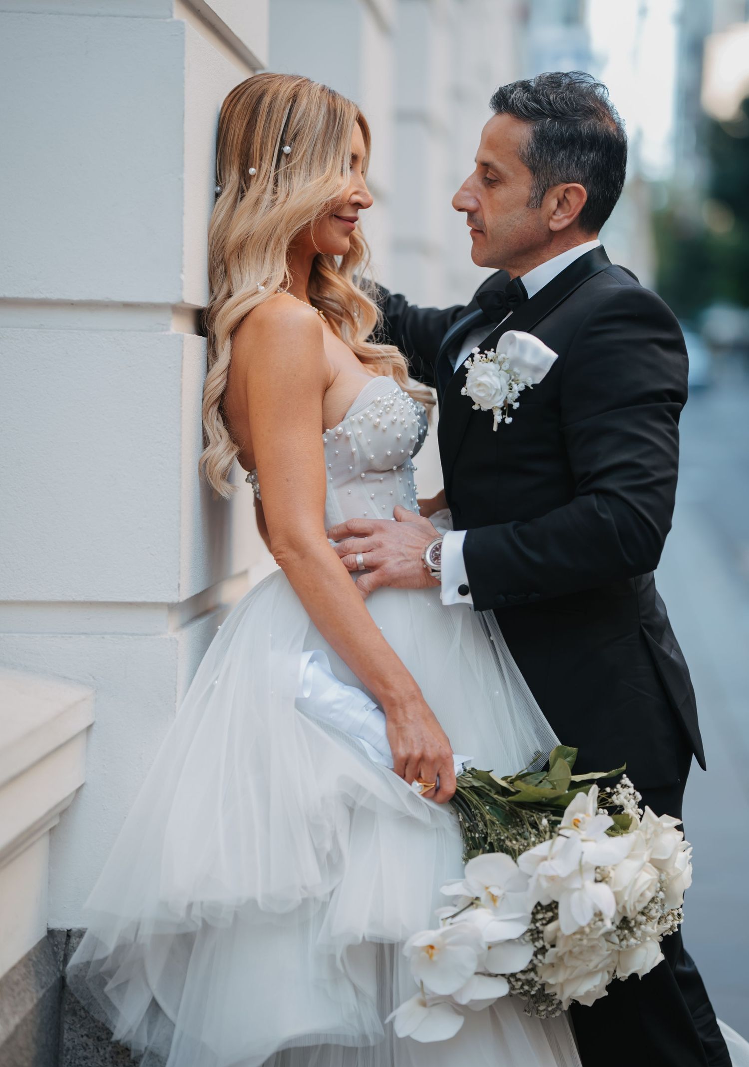 A couple in wedding attire share an intimate moment against a white pillar during their outdoor photoshoot.