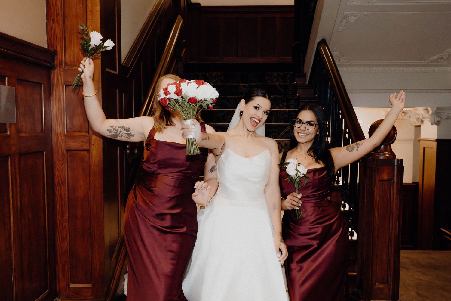 Bride in white gown poses with two bridesmaids in burgundy satin dresses holding bouquets on elegant staircase.