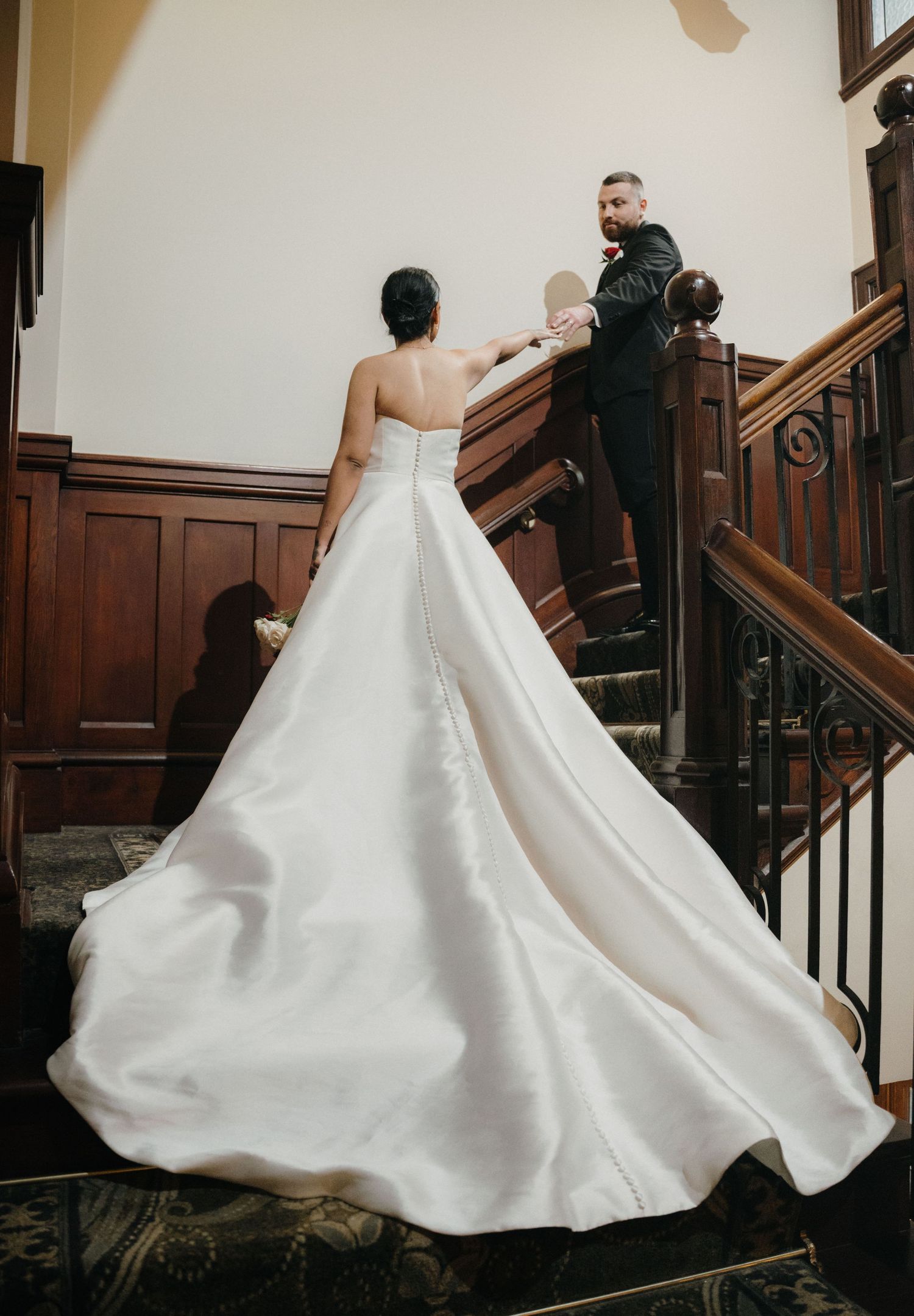 Bride in a flowing white strapless ball gown on wooden staircase with groom in black suit during first look.