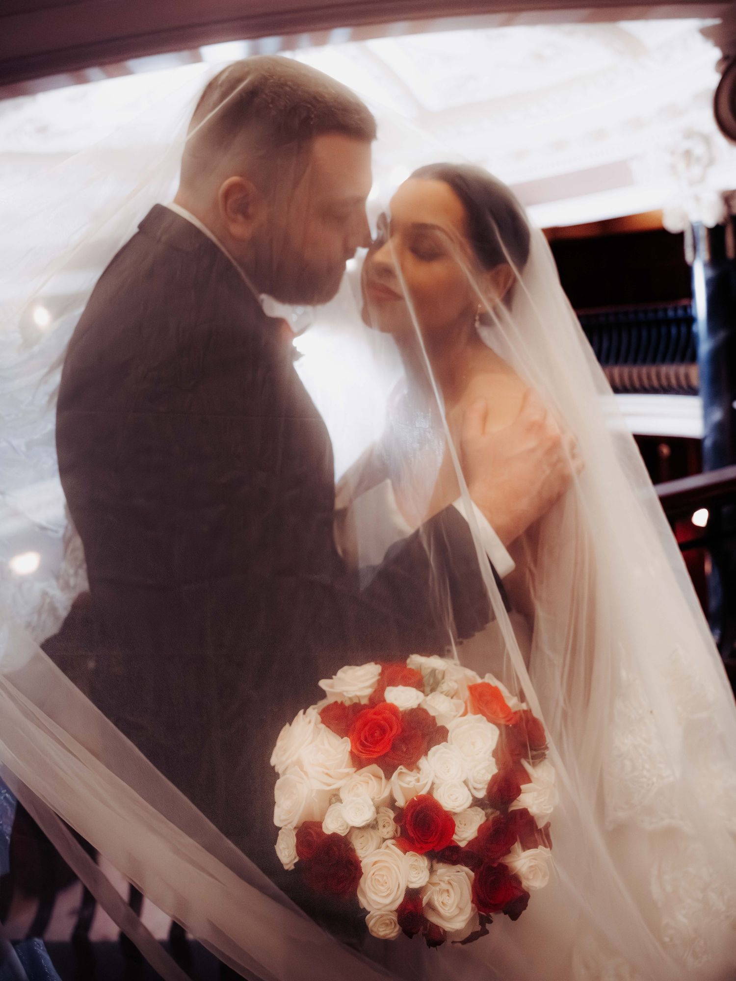 Romantic wedding couple sharing an intimate moment under a veil, bride holding red and white rose bouquet.