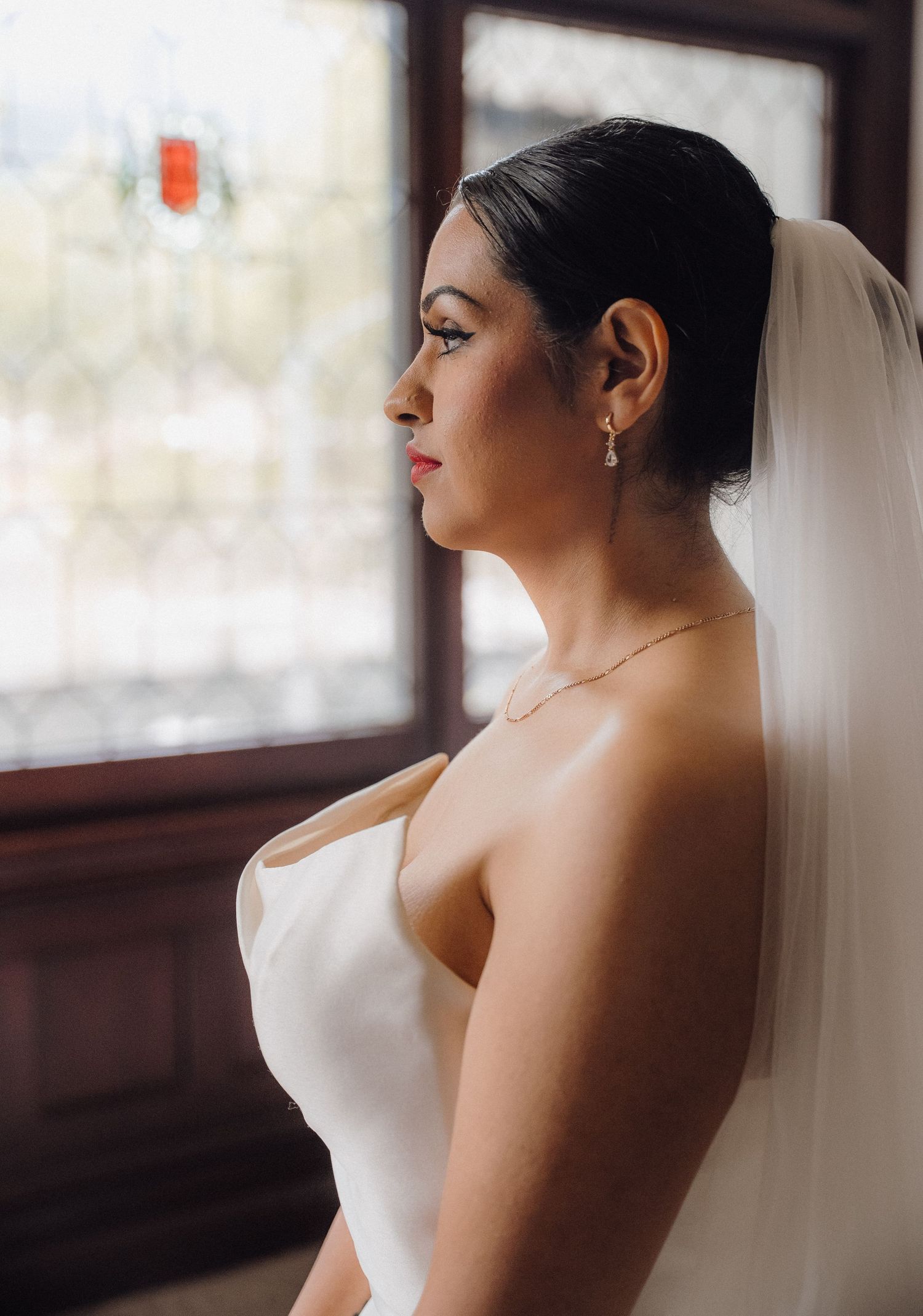 Elegant bride in strapless white gown and veil gazes out window, wearing pearl earrings and delicate necklace.