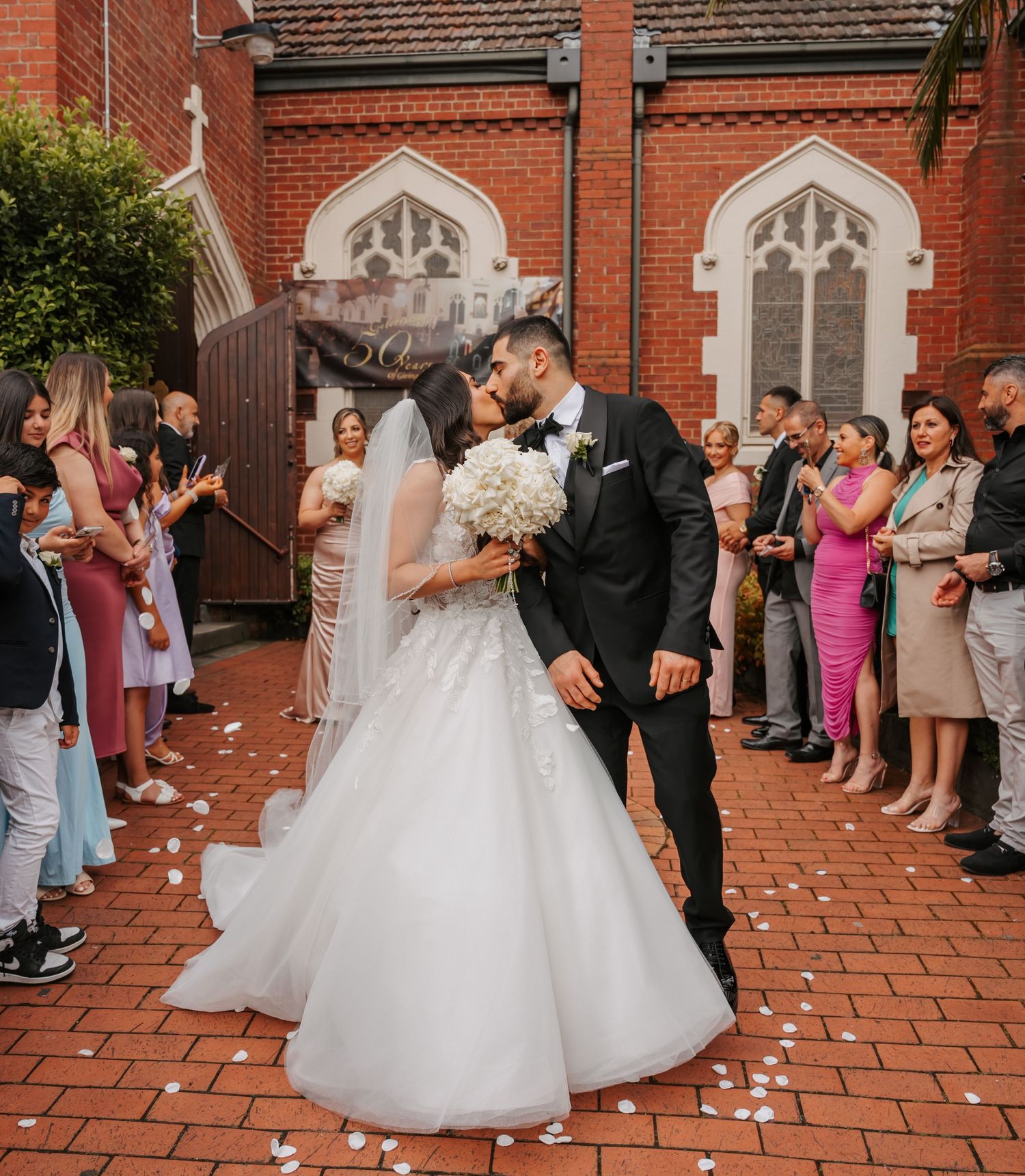 Wedding couple shares a kiss on brick patio outside church as guests shower them with flower petals in celebration.
