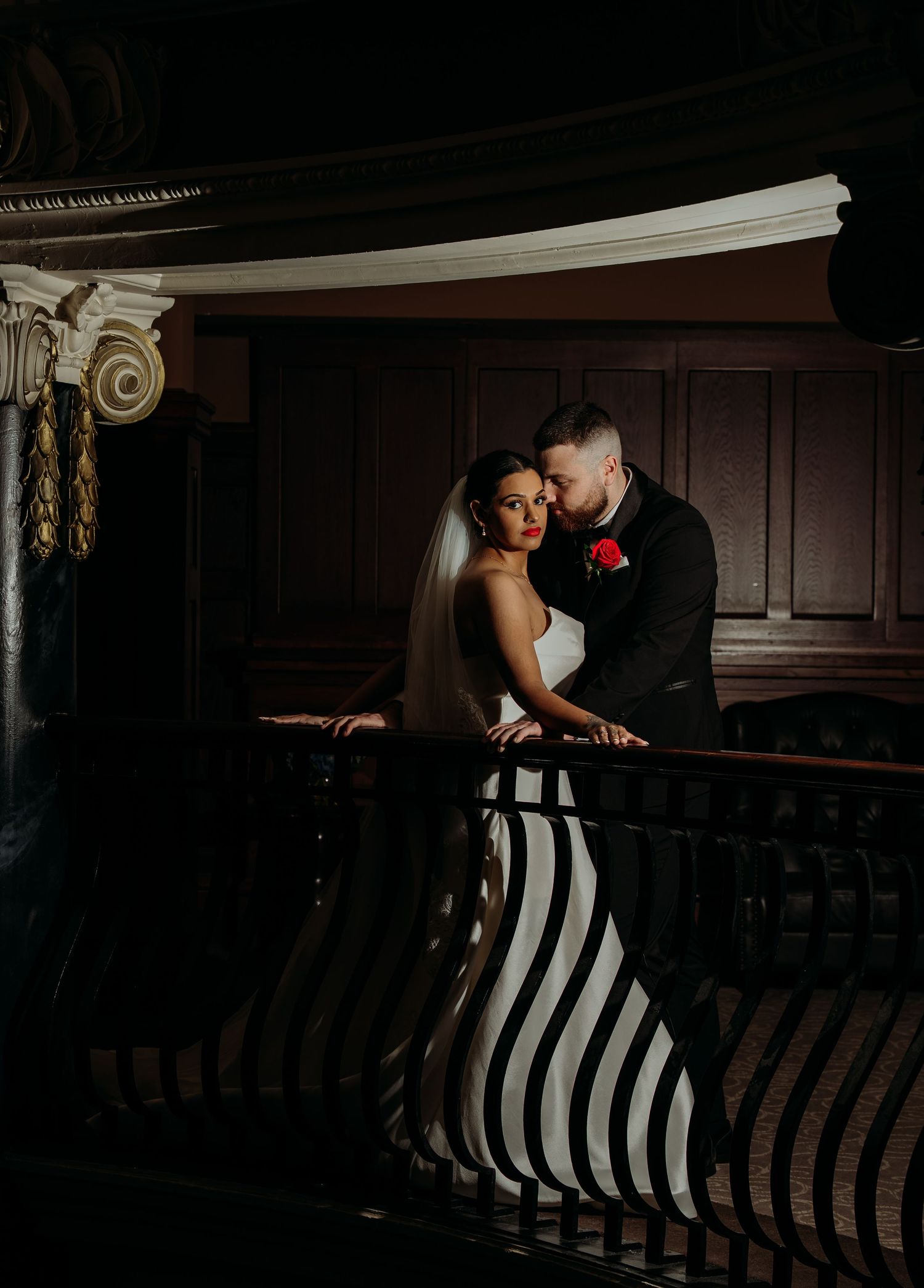 Bride and groom share a romantic embrace on a grand staircase balcony in an elegant dark wedding venue.