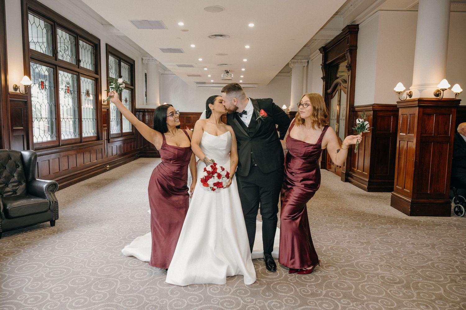 Bride and groom share a kiss while two bridesmaids in burgundy satin dresses celebrate beside them in elegant venue.