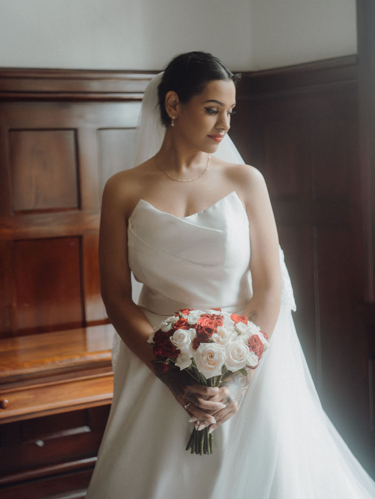 Bride in strapless white gown holds red and white rose bouquet, standing by wooden paneling in soft natural light.