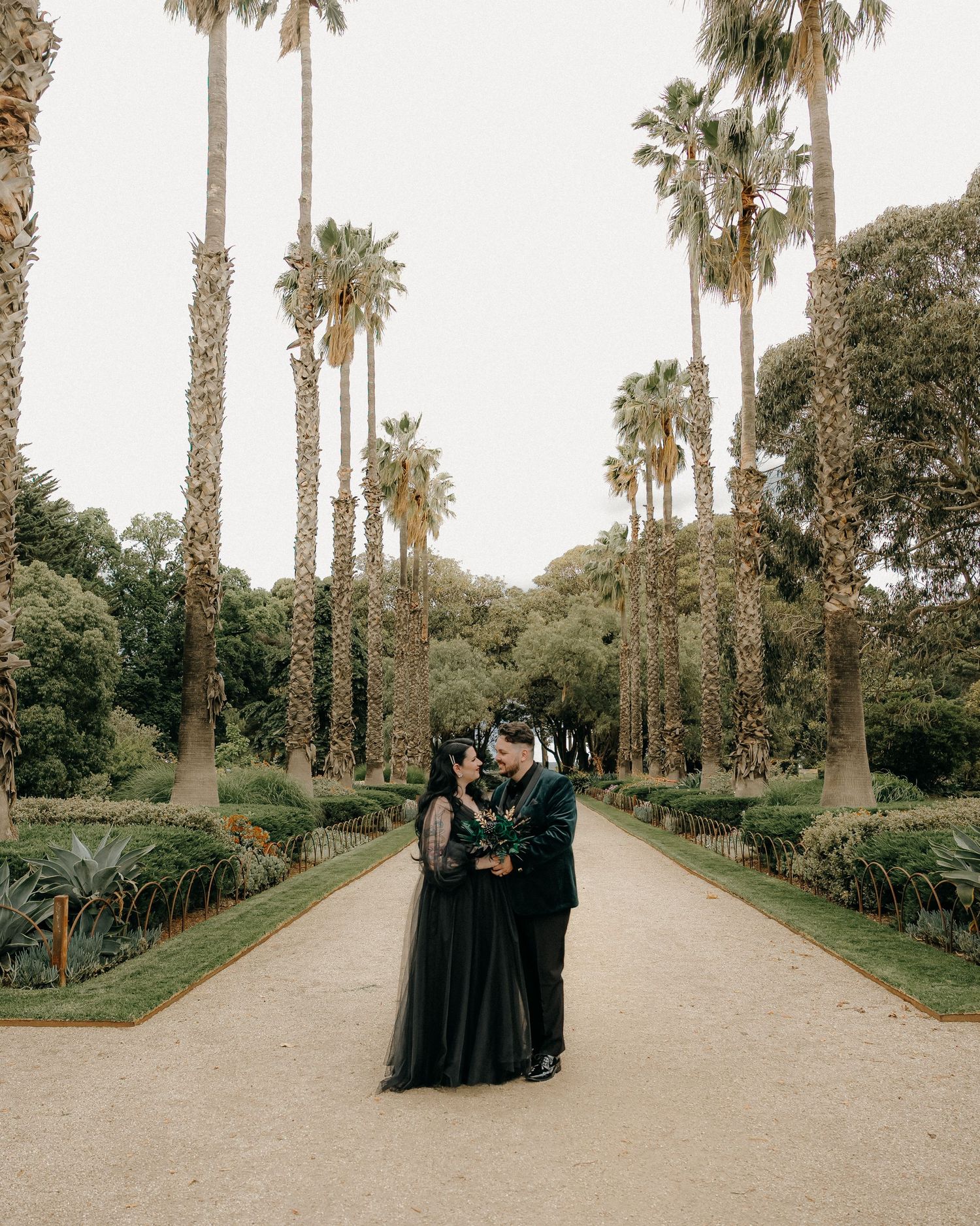 A romantic palm tree-lined pathway in a lush garden setting with figures in black attire walking along the path.