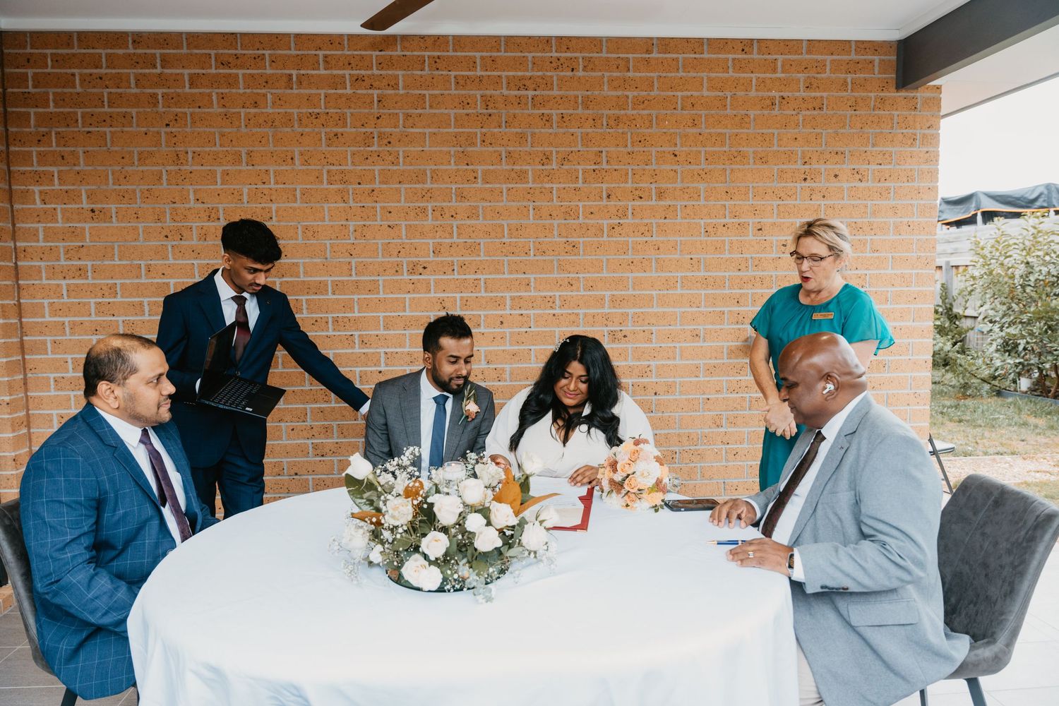 Wedding party gathers in front of a brick wall, with elegant floral arrangements and formal attire.