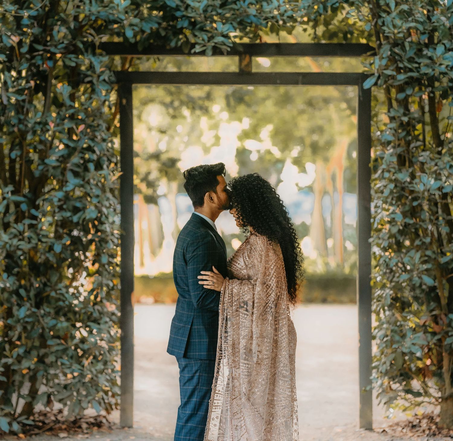 Romantic couple embraces under a lush garden archway wearing elegant navy suit and sequined gown at sunset.