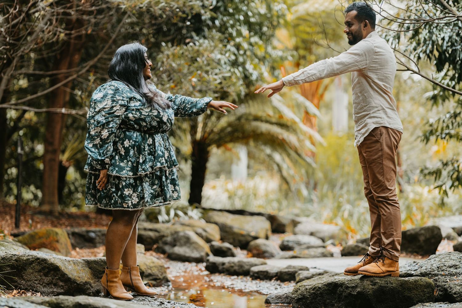 Two people helping each other cross rocks in a garden path surrounded by lush foliage and trees.