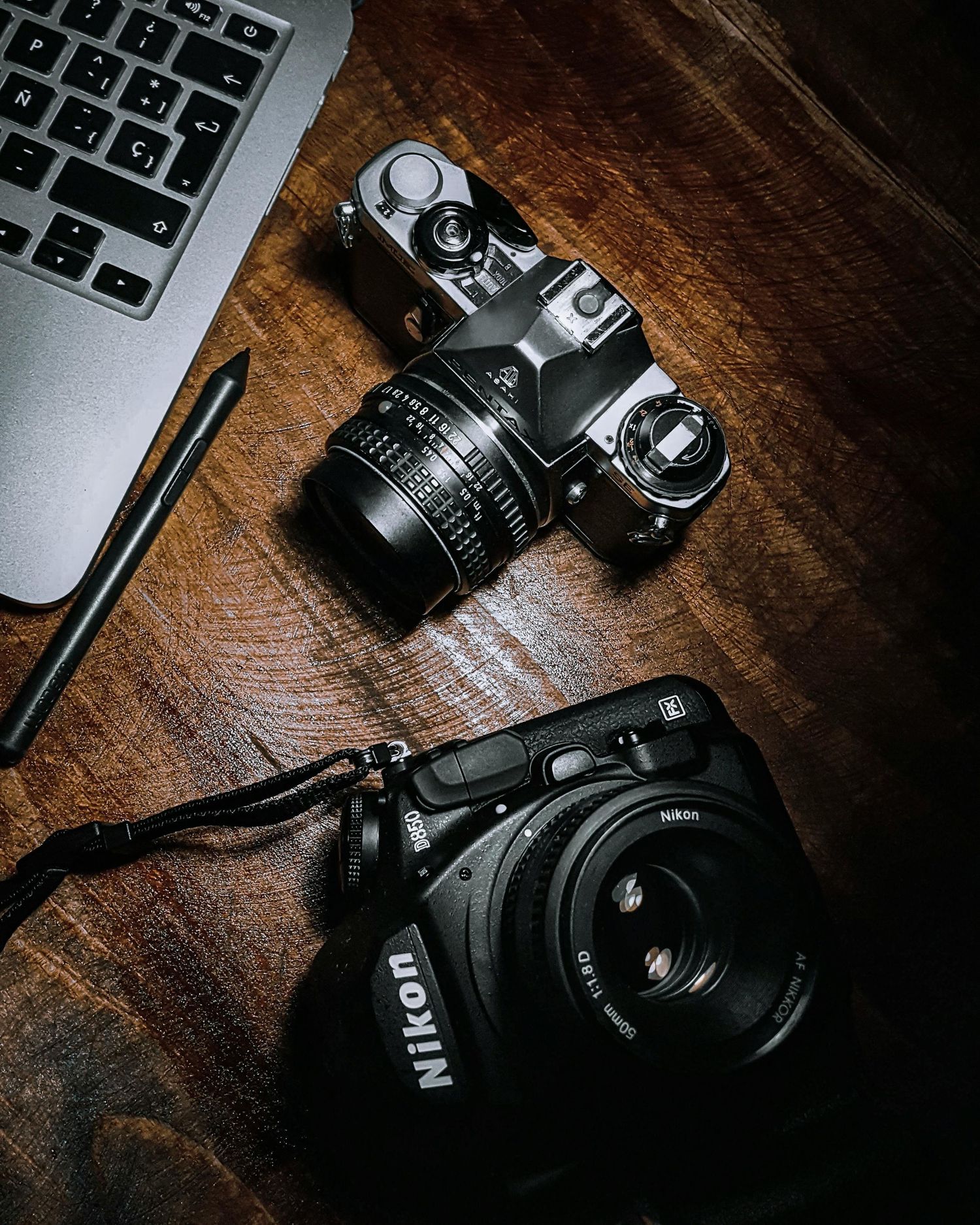 A black DSLR camera sits next to a laptop keyboard on a dark wooden surface in moody lighting.